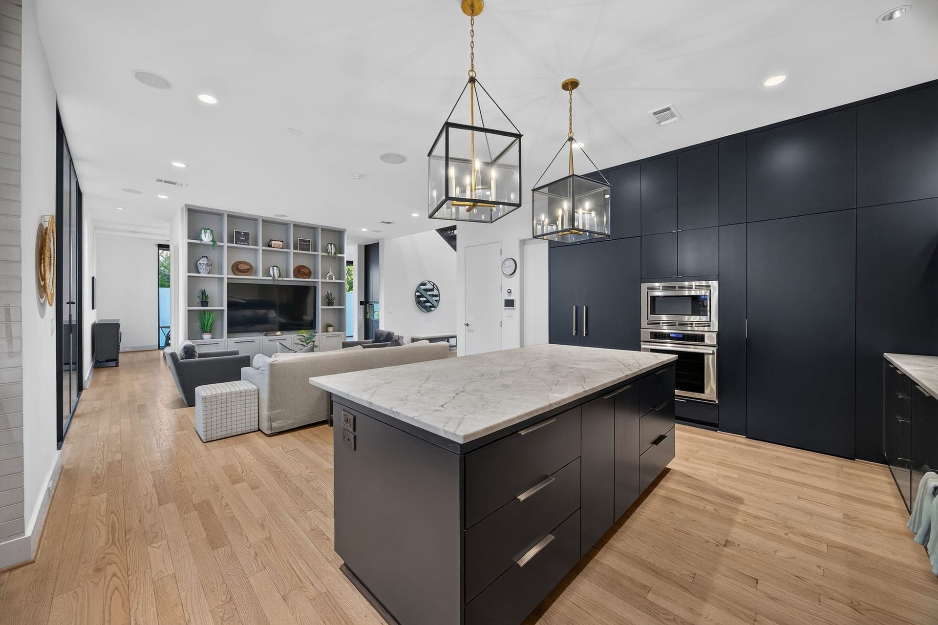 6002 Valley Forge Drive, Houston, TX 77057 Sleek modern kitchen with a large island and elegant pendant lighting, seamlessly flowing into a spacious living area with built-in shelving and hardwood floors.