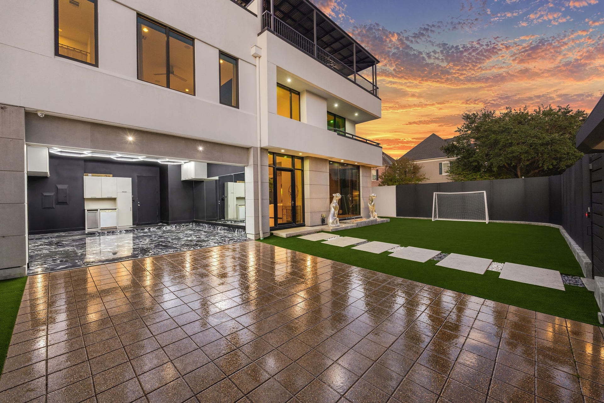 6002 Valley Forge Drive, Houston, TX 77057 Experience contemporary elegance with this spacious outdoor area featuring a sleek patio, lush artificial turf, and a versatile garage space, perfect for entertaining under stunning sunset skies.