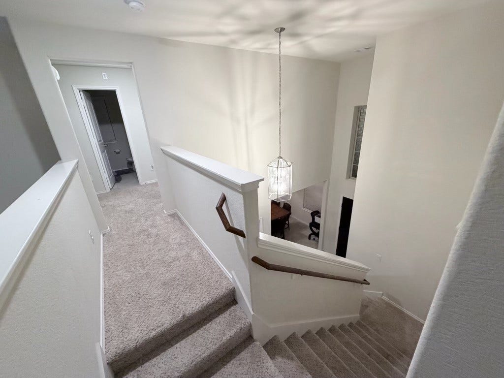 2806 Bretton Woods Drive, Conroe, TX 77301 This photo shows a carpeted staircase with a wooden handrail leading down to a dining area. The space is well-lit by a modern hanging light fixture, and there's a hallway with a visible doorway on the upper level. The walls are painted a neutral color, enhancing the bright and open feel.