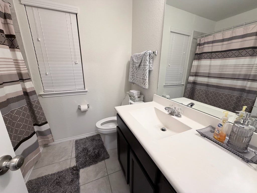 2806 Bretton Woods Drive, Conroe, TX 77301 This bathroom features a clean and modern design with neutral tones. It includes a shower-tub combo with a striped curtain, a spacious vanity with a large mirror, and tiled flooring. The window provides natural light, and there are tasteful accessories for a cozy feel.