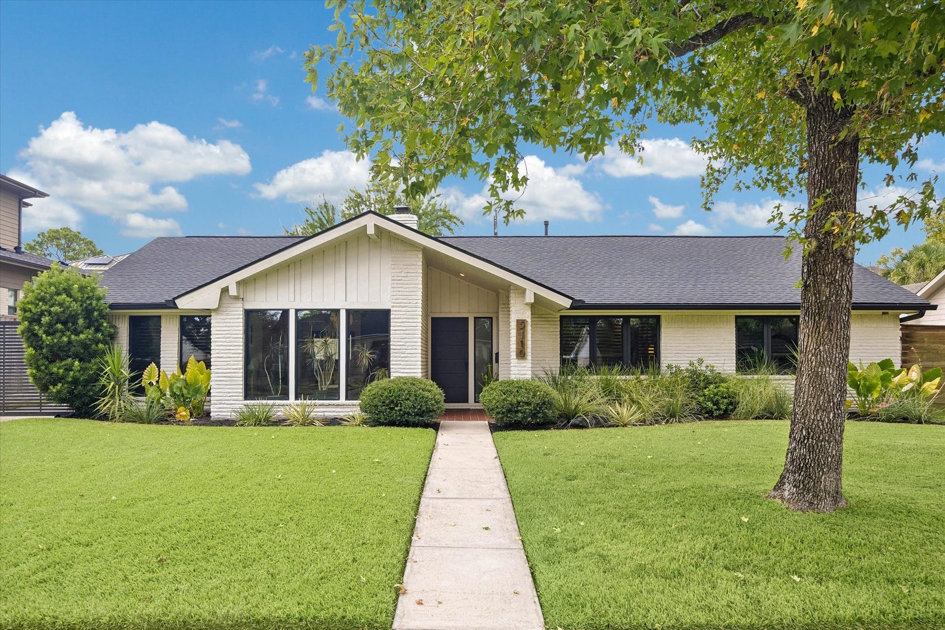 Stunning curb appeal showcases this thoughtfully renovated home, featuring a white painted brick exterior, expansive picture windows, lush landscaping, and a welcoming front entry.