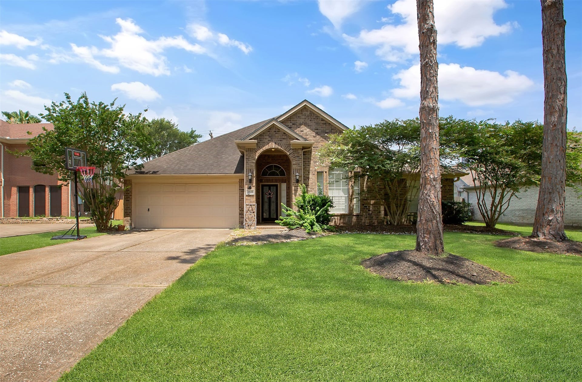 This charming single-story brick home features a neatly landscaped front yard with mature trees and a wide driveway leading to a two-car garage. Perfect for family living in a serene neighborhood.