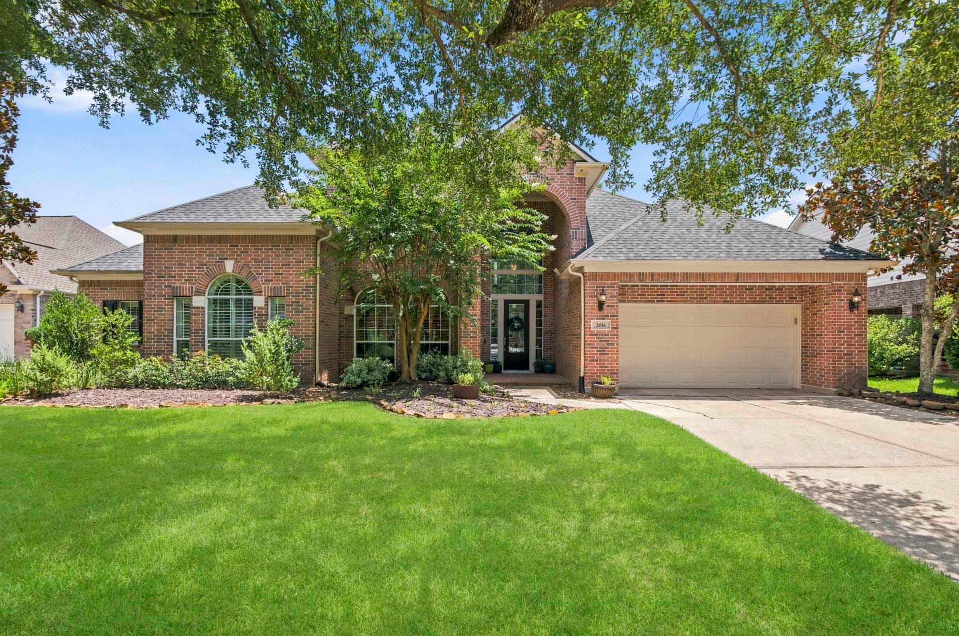 Charming brick home with a spacious three-car garage and lush front lawn, framed by mature trees and vibrant landscaping.