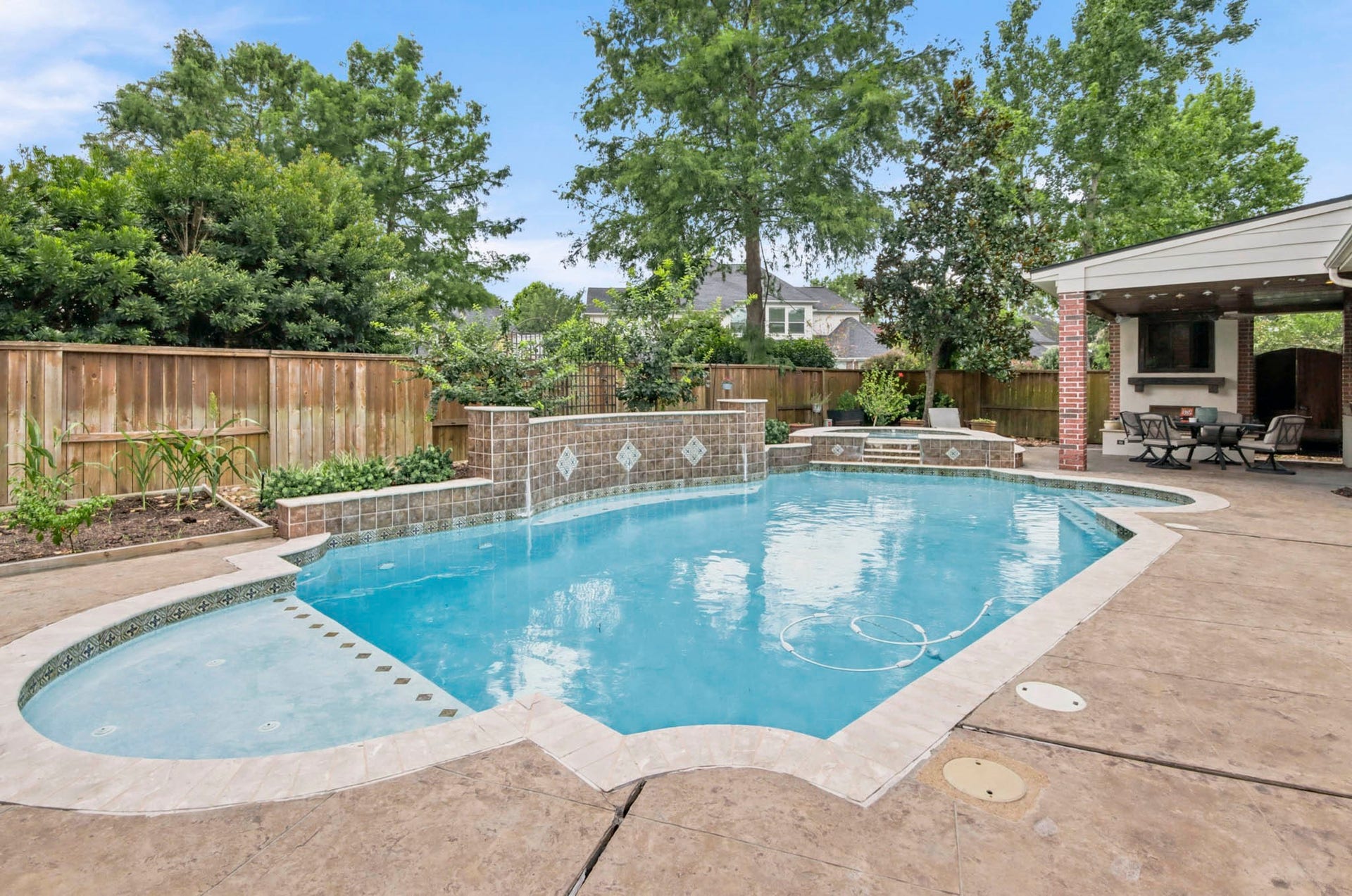 A spacious backyard oasis featuring a sparkling pool with a charming waterfall feature, surrounded by lush greenery and a covered patio perfect for outdoor dining and relaxation.
