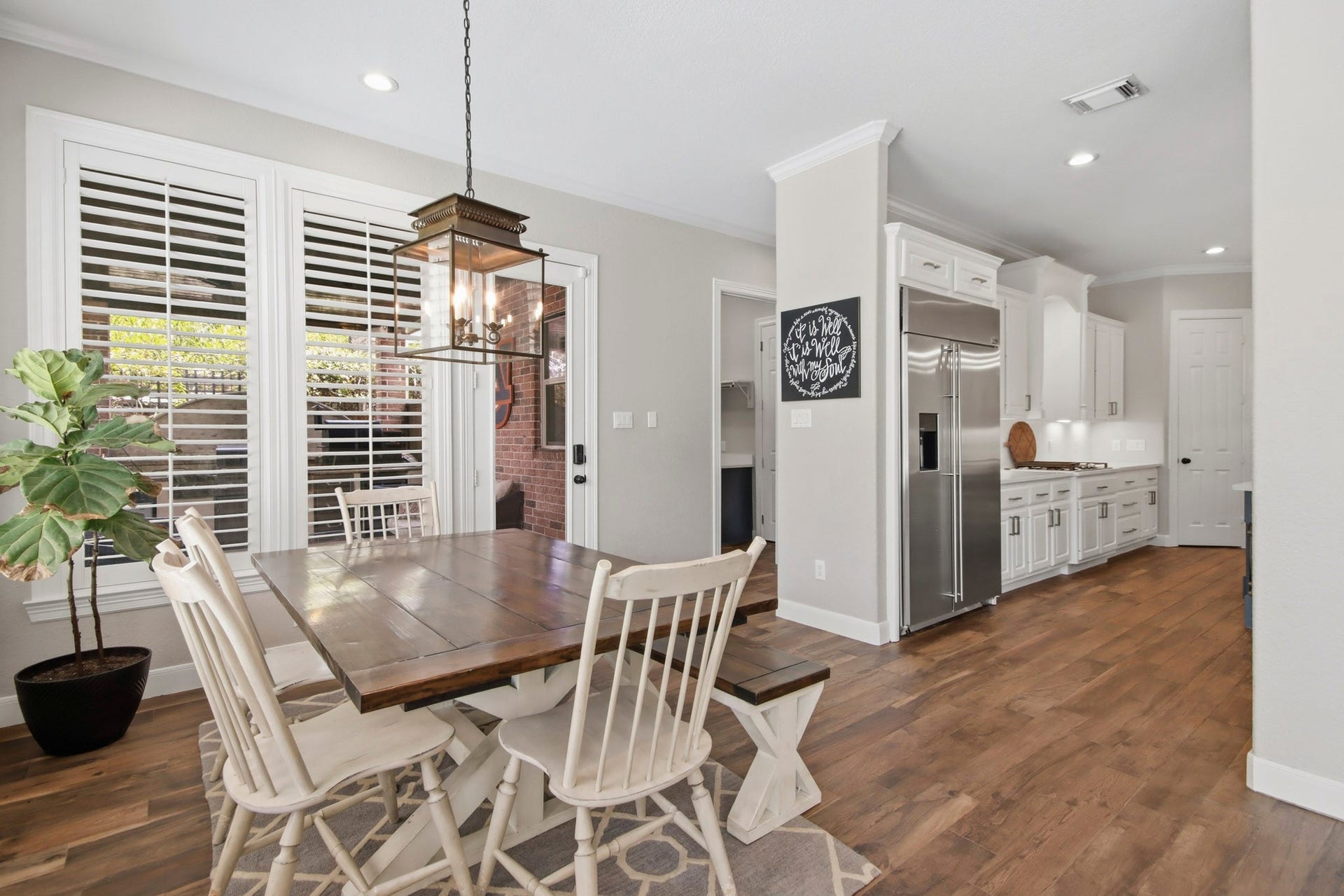 Bright and inviting breakfast area with elegant light fixture, and large windows with shutters, seamlessly connected to kitchen and living room.