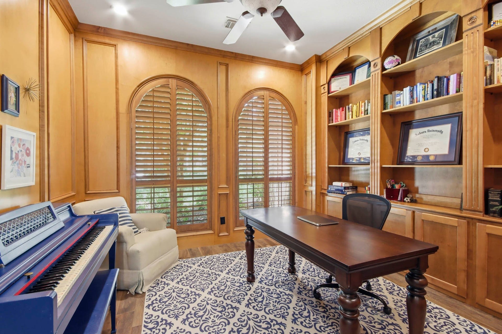 Home office featuring rich wood paneling, custom built-in bookshelves, and arched windows with shutters creating a perfect space for productivity and inspiration.