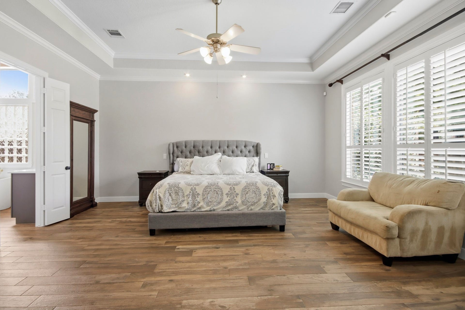 Spacious bedroom with wood-like tile floors, ample natural light streaming through large shuttered windows, complemented by enough space for a cozy seating area.