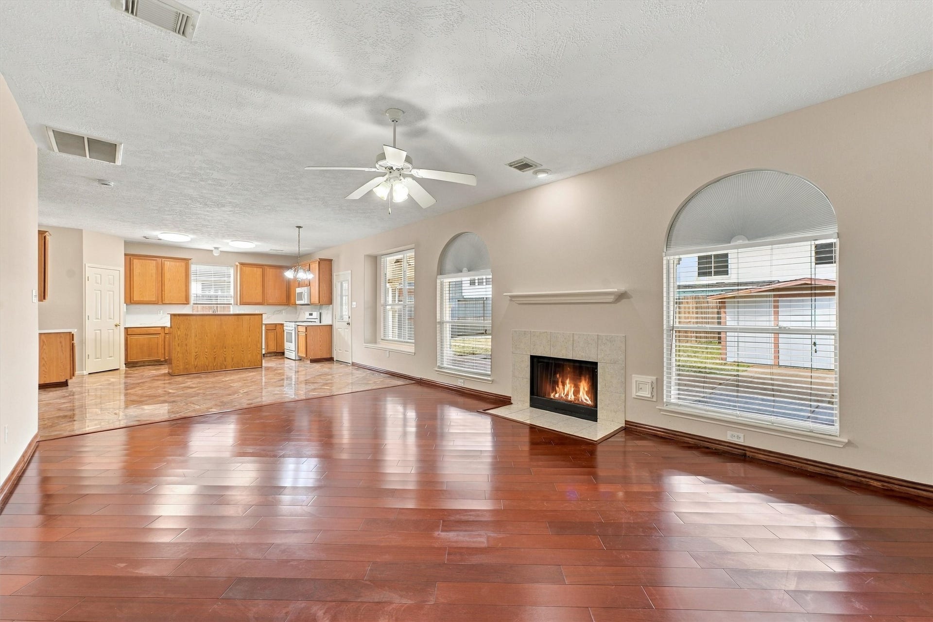 Spacious open-plan living area featuring rich hardwood floors, a cozy fireplace, and large windows offering plenty of natural light, seamlessly flowing into a modern kitchen with ample cabinetry and a center island.