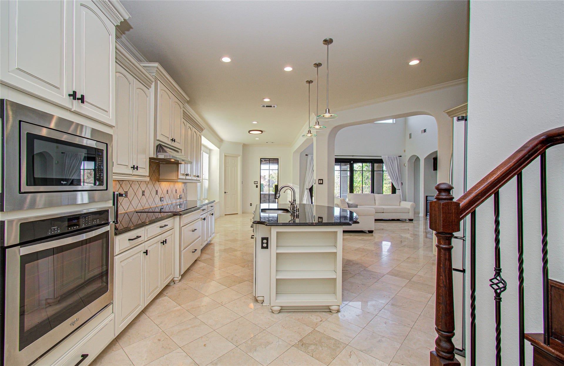 This photo showcases a spacious, elegant kitchen with white cabinetry, stainless steel appliances, and a central island with a dark countertop. The open layout leads into a bright living area with large windows and light tile flooring, creating a seamless flow between spaces.
