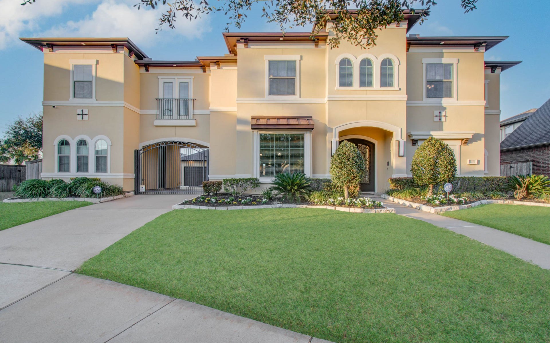 This is a spacious, two-story home with a stucco exterior and elegant architectural details. It features a well-maintained lawn, landscaped garden, and a gated driveway. The house has large windows, offering plenty of natural light.