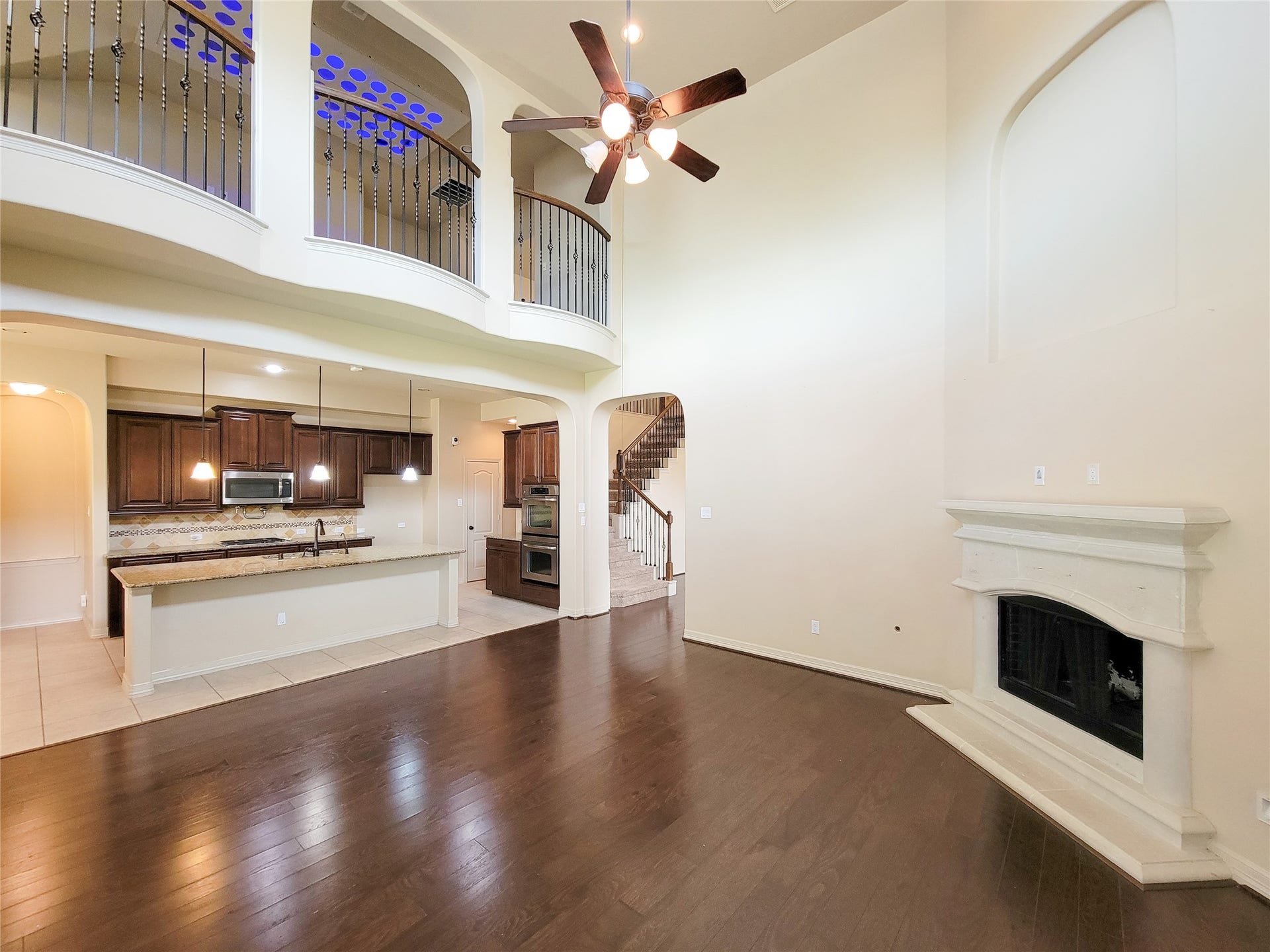 2210 Pearl Bay Court, Pearland, TX 77584 This spacious living area features high ceilings, a modern ceiling fan, and elegant dark wood flooring. The open layout seamlessly connects to a kitchen with granite countertops, stainless steel appliances, and warm wood cabinetry. A cozy fireplace adds charm, and a second-floor balcony overlooks the room, enhancing its airy feel.
