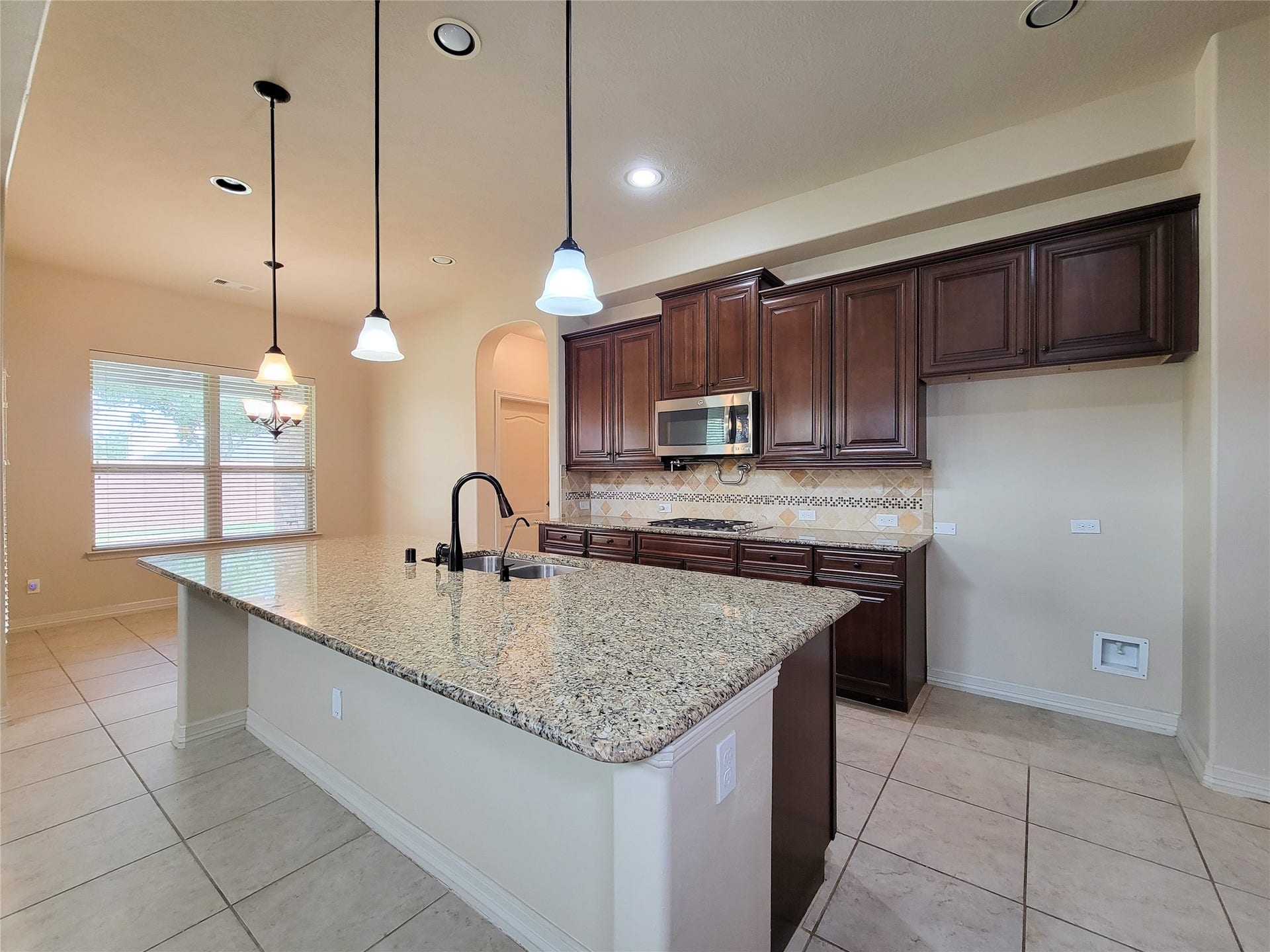 2210 Pearl Bay Court, Pearland, TX 77584 This kitchen features a spacious granite island with a sink, modern pendant lighting, and ample dark wood cabinetry. It includes a built-in microwave and stylish tile backsplash, with large windows providing natural light.