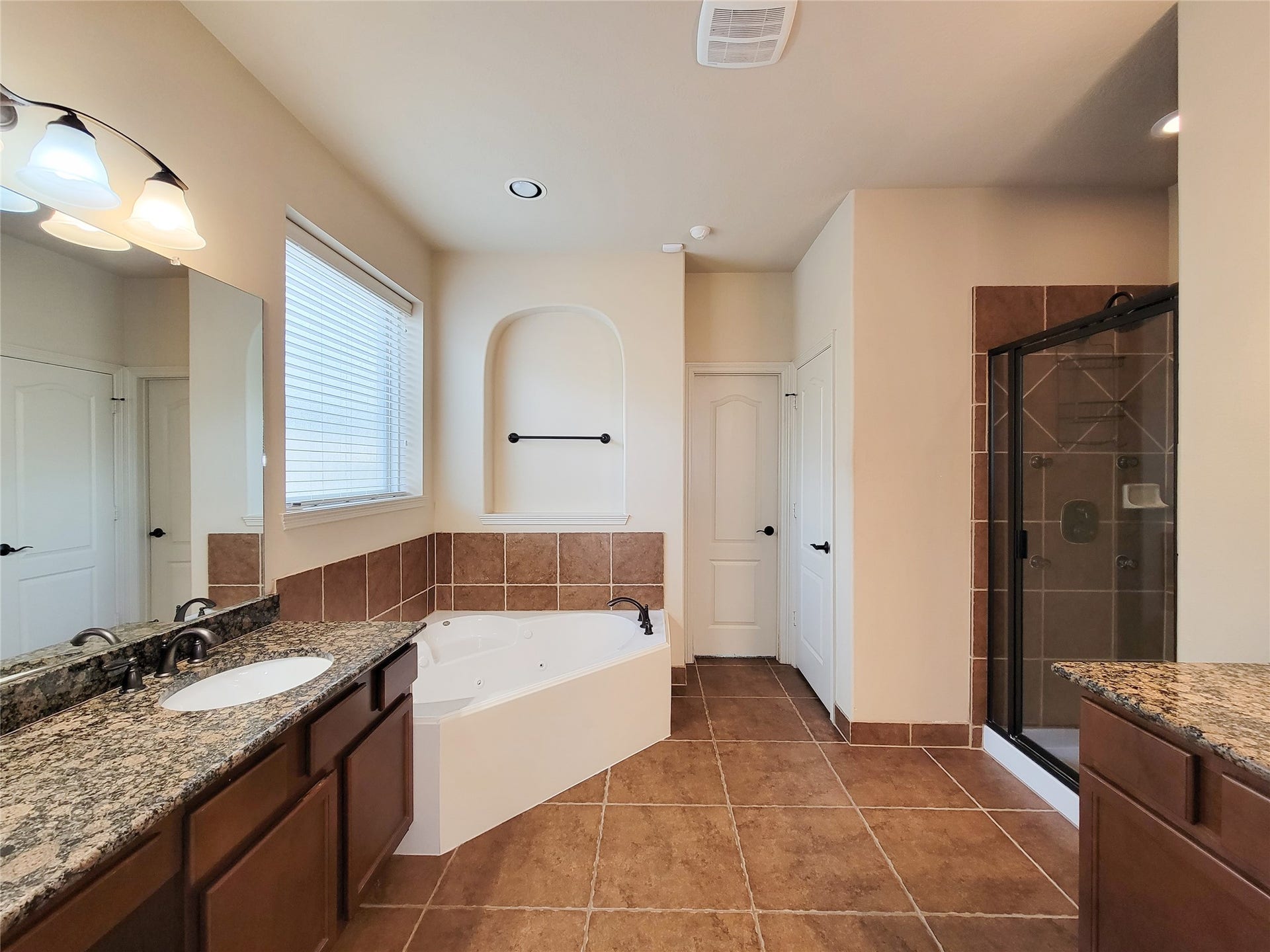 2210 Pearl Bay Court, Pearland, TX 77584 This spacious bathroom features a luxurious soaking tub, a separate glass-enclosed shower, dual granite-topped vanities, and warm tile flooring. Natural light streams in through a large window, creating a bright and inviting atmosphere.