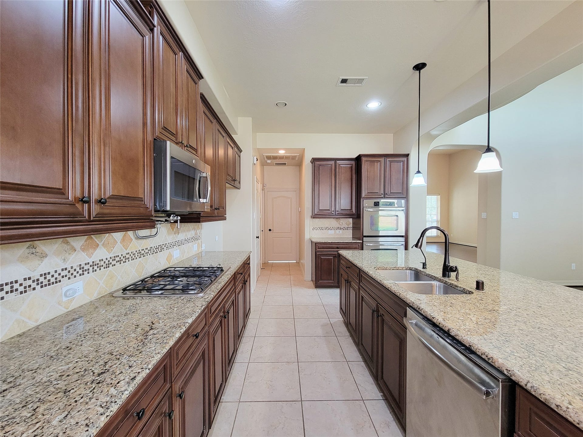 2210 Pearl Bay Court, Pearland, TX 77584 This kitchen features elegant wooden cabinetry, granite countertops, and stainless steel appliances. It includes a built-in oven, microwave, gas cooktop, and a spacious island with a double sink and dishwasher. The tile backsplash and pendant lighting add a touch of sophistication.