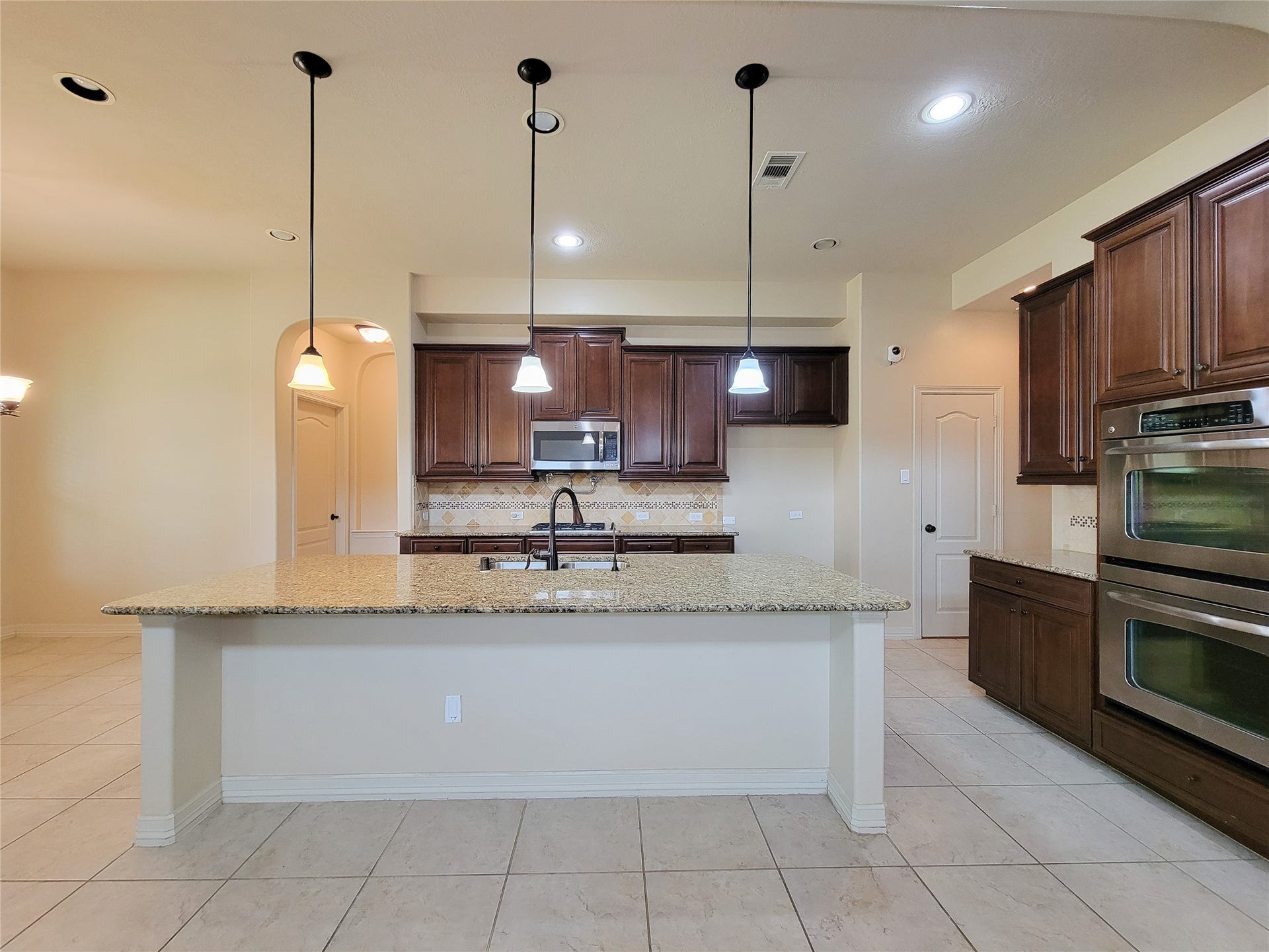 2210 Pearl Bay Court, Pearland, TX 77584 This kitchen features a spacious island with a granite countertop, pendant lighting, and stainless steel appliances. It has ample cabinet space in rich wood tones, a double oven, and a tiled floor, creating a warm and functional cooking area.