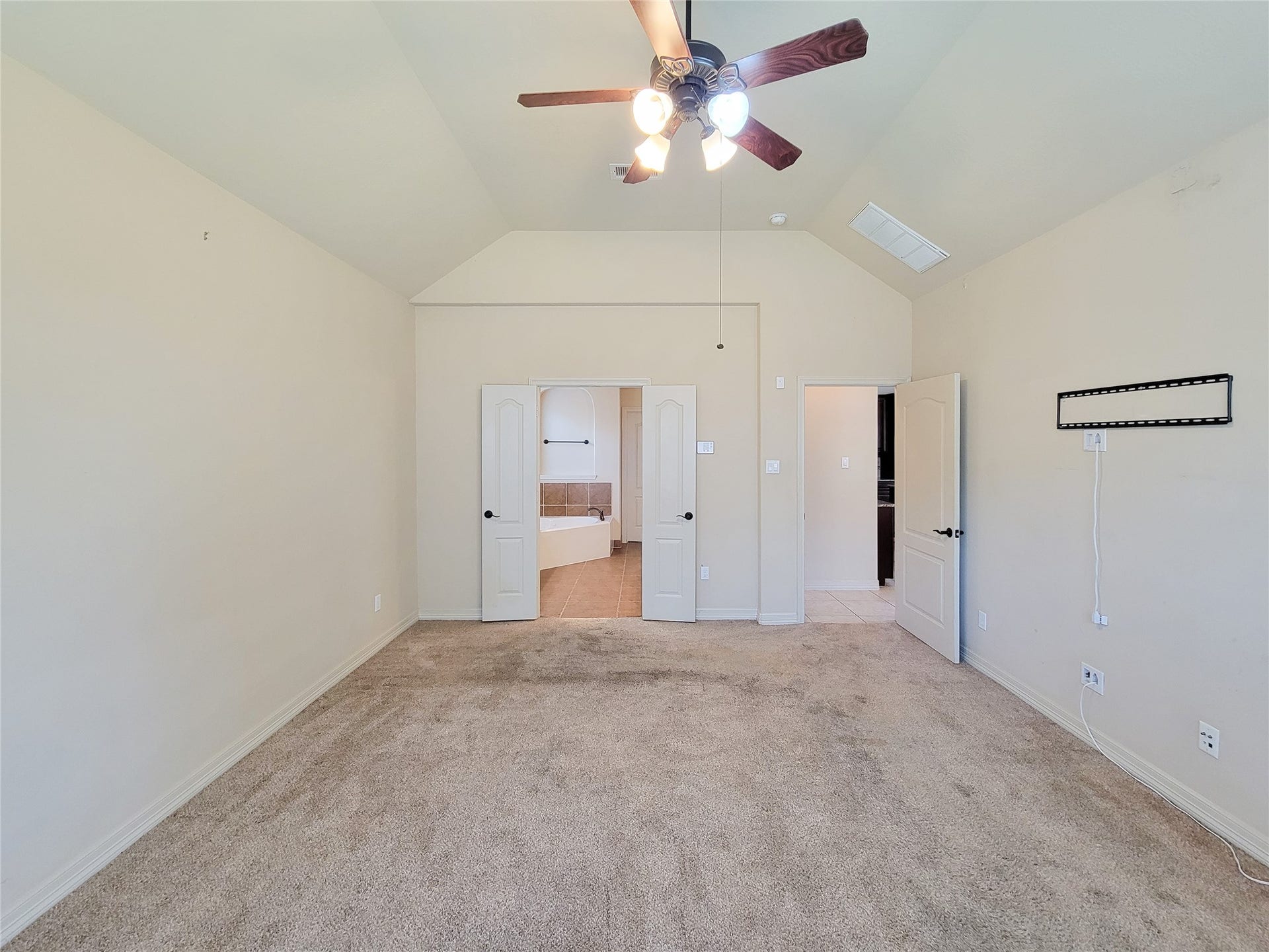 2210 Pearl Bay Court, Pearland, TX 77584 This spacious, carpeted bedroom features neutral walls, a ceiling fan, and double doors leading to a bathroom with a bathtub. There's ample natural light and a wall-mounted bracket for a TV.