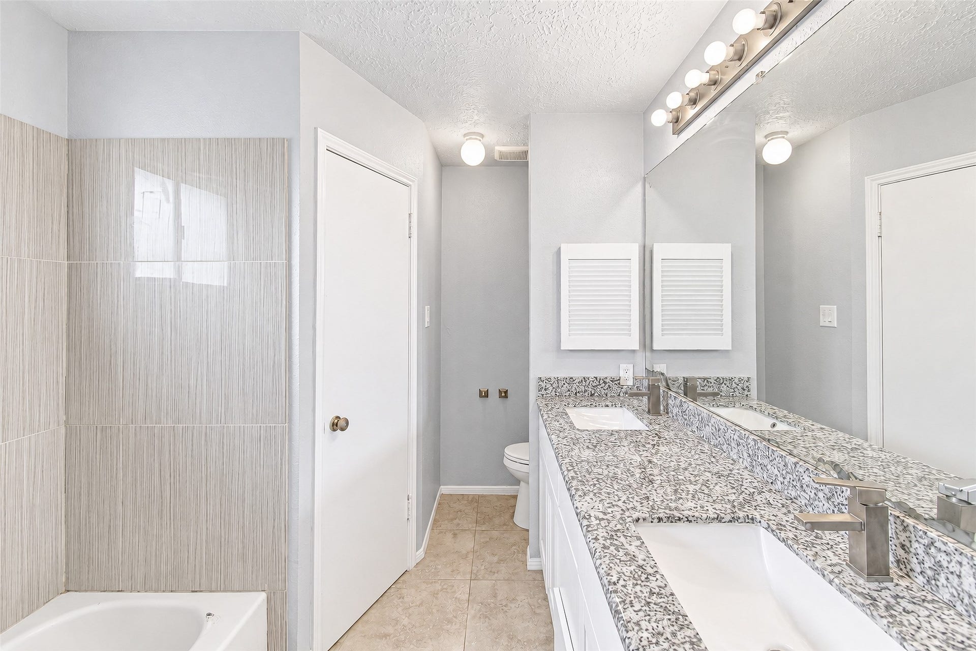 12027 Huntington Park Drive, Houston, TX 77099 Bright and modern bathroom featuring dual sinks with sleek granite countertops, a large mirror, and stylish light fixtures. Neutral tile surrounds the bathtub, creating a clean and inviting atmosphere.