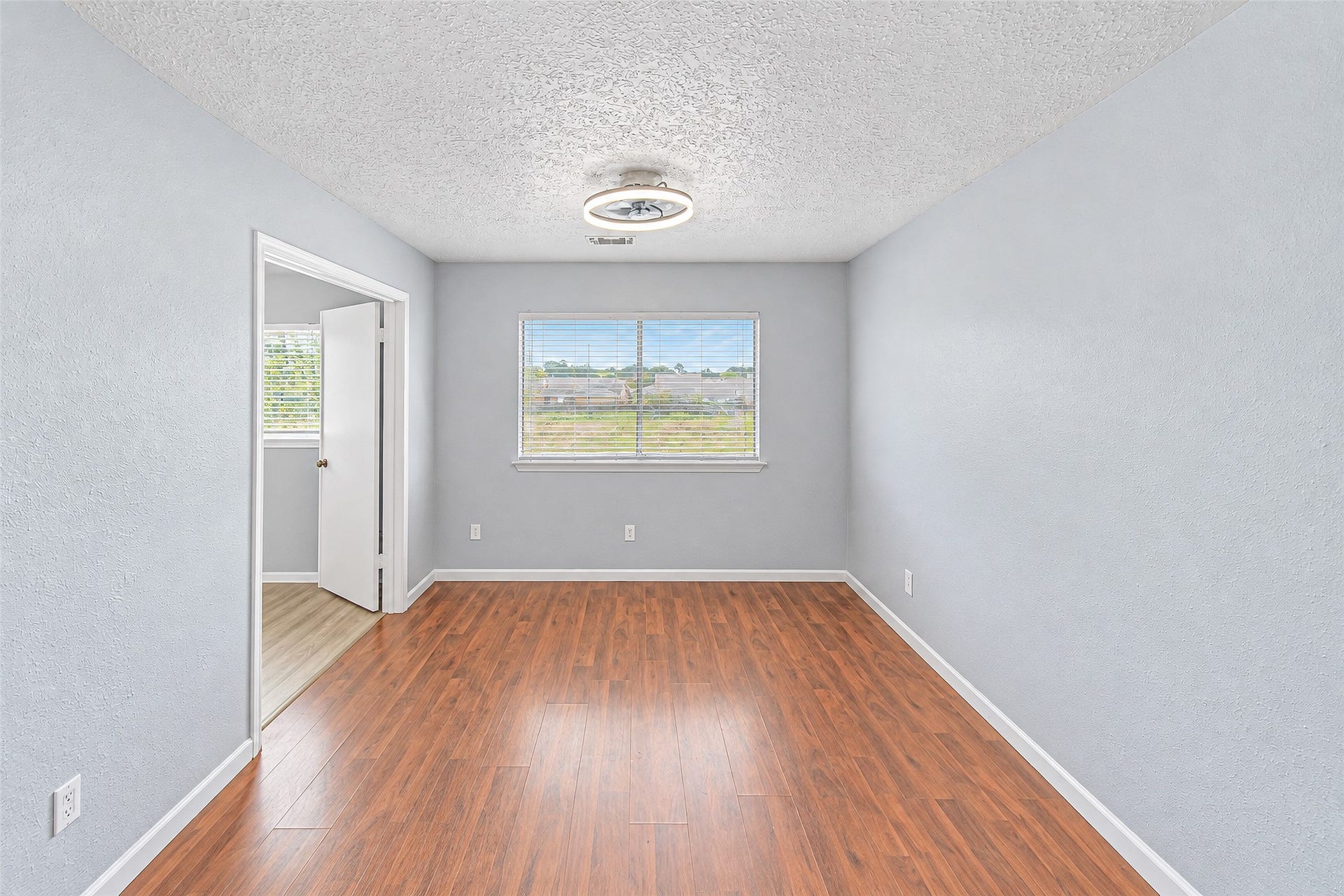 12027 Huntington Park Drive, Houston, TX 77099 Bright and inviting room featuring sleek hardwood floors, soft gray walls, and a modern light fixture. Abundant natural light flows through the window, highlighting the open layout and creating a warm atmosphere.