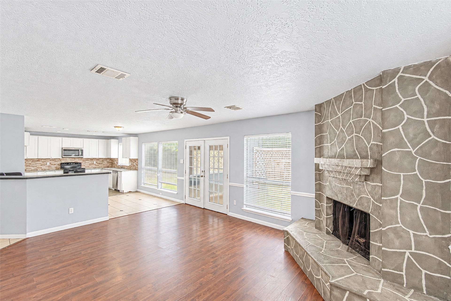 12027 Huntington Park Drive, Houston, TX 77099 Spacious living area with elegant hardwood floors, a unique stone-patterned fireplace, and abundant natural light from large windows and French doors. The open-concept layout flows seamlessly into a modern kitchen with sleek cabinetry and stainless steel appliances.