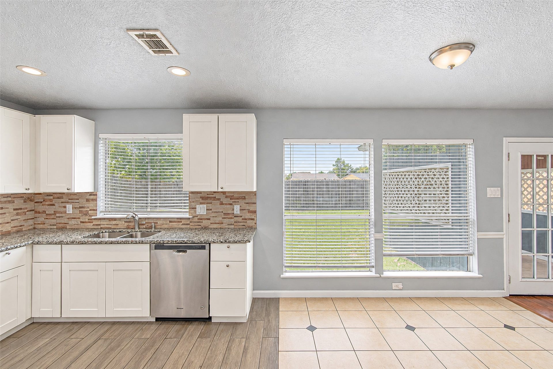 12027 Huntington Park Drive, Houston, TX 77099 Bright kitchen and dining area featuring modern white cabinets, granite countertops, and a stylish tile backsplash, with ample natural light from large windows overlooking a spacious backyard.