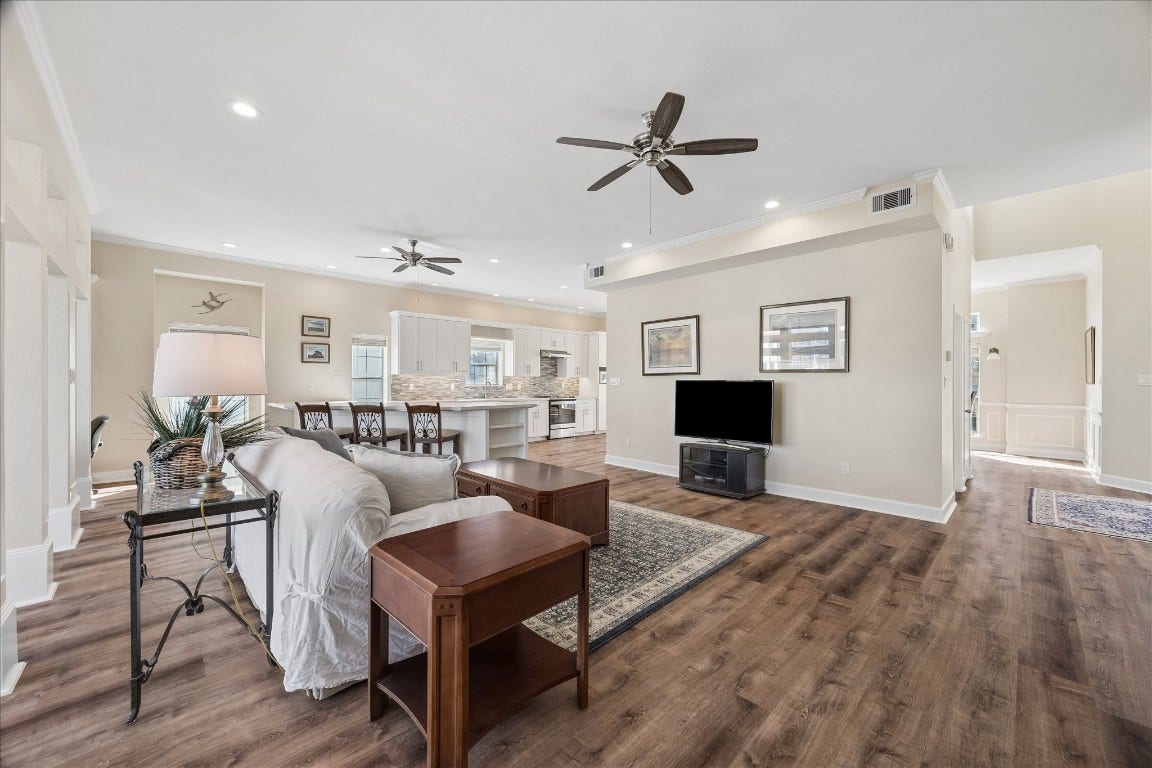 This open-concept living area features stylish hardwood flooring, neutral walls, and recessed lighting. It includes a spacious living room with ceiling fans, a cozy sofa, and a TV setup. The adjacent kitchen boasts a large island, modern appliances, and white cabinetry, offering a seamless flow for entertaining.