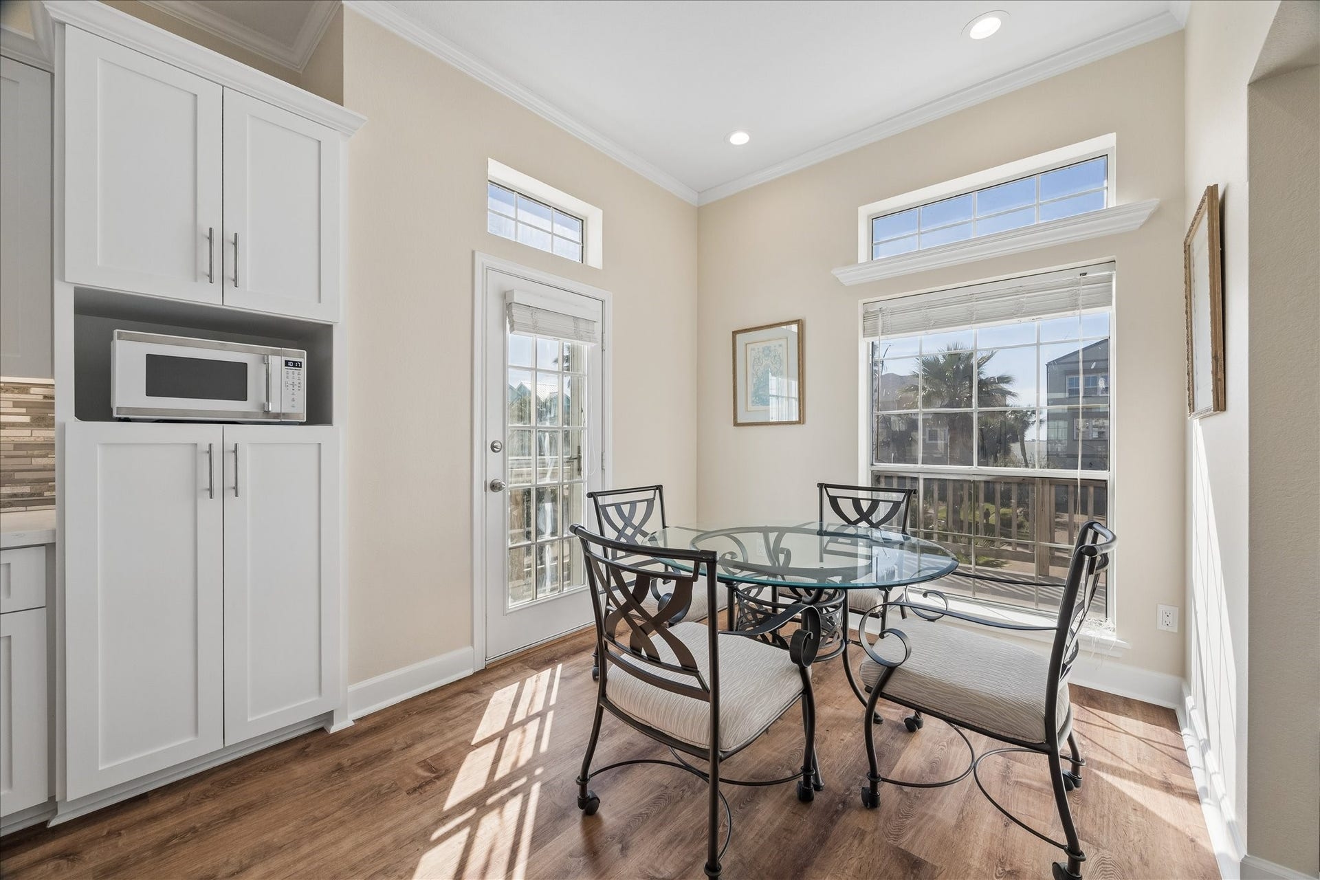 This bright dining area features large windows, a glass-top table with elegant chairs, and access to a patio through a door with glass panels. The adjacent cabinetry provides storage and includes a built-in microwave. The space is enhanced by natural light and wood flooring.
