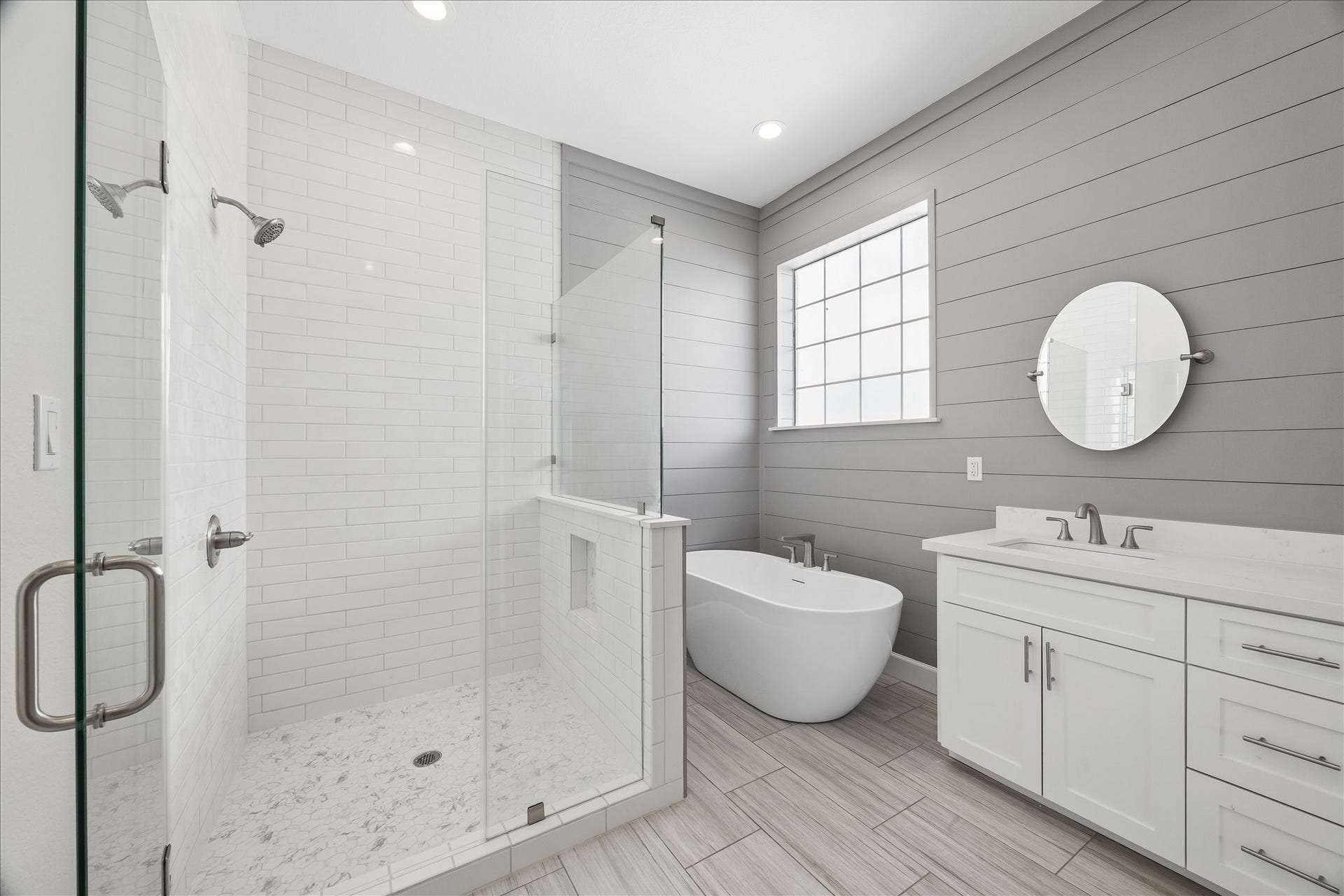 This bathroom features a modern design with a spacious glass-enclosed shower, a freestanding bathtub, and a double vanity with sleek fixtures. The room is brightened by a large window and has elegant gray shiplap walls and light wood flooring.