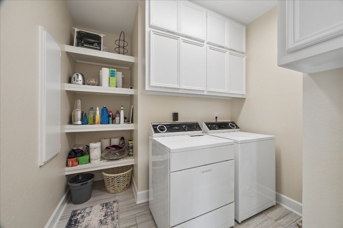 This photo showcases a well-organized laundry room featuring a washer and dryer, ample cabinetry for storage, and open shelving for easy access to supplies. The room has a clean, modern look with neutral walls and flooring.