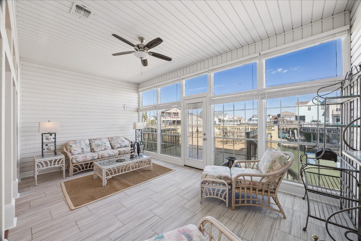 This bright sunroom features large windows with waterfront views, white wood-panel walls, and a cozy seating area with wicker furniture, perfect for relaxing and enjoying the scenery.