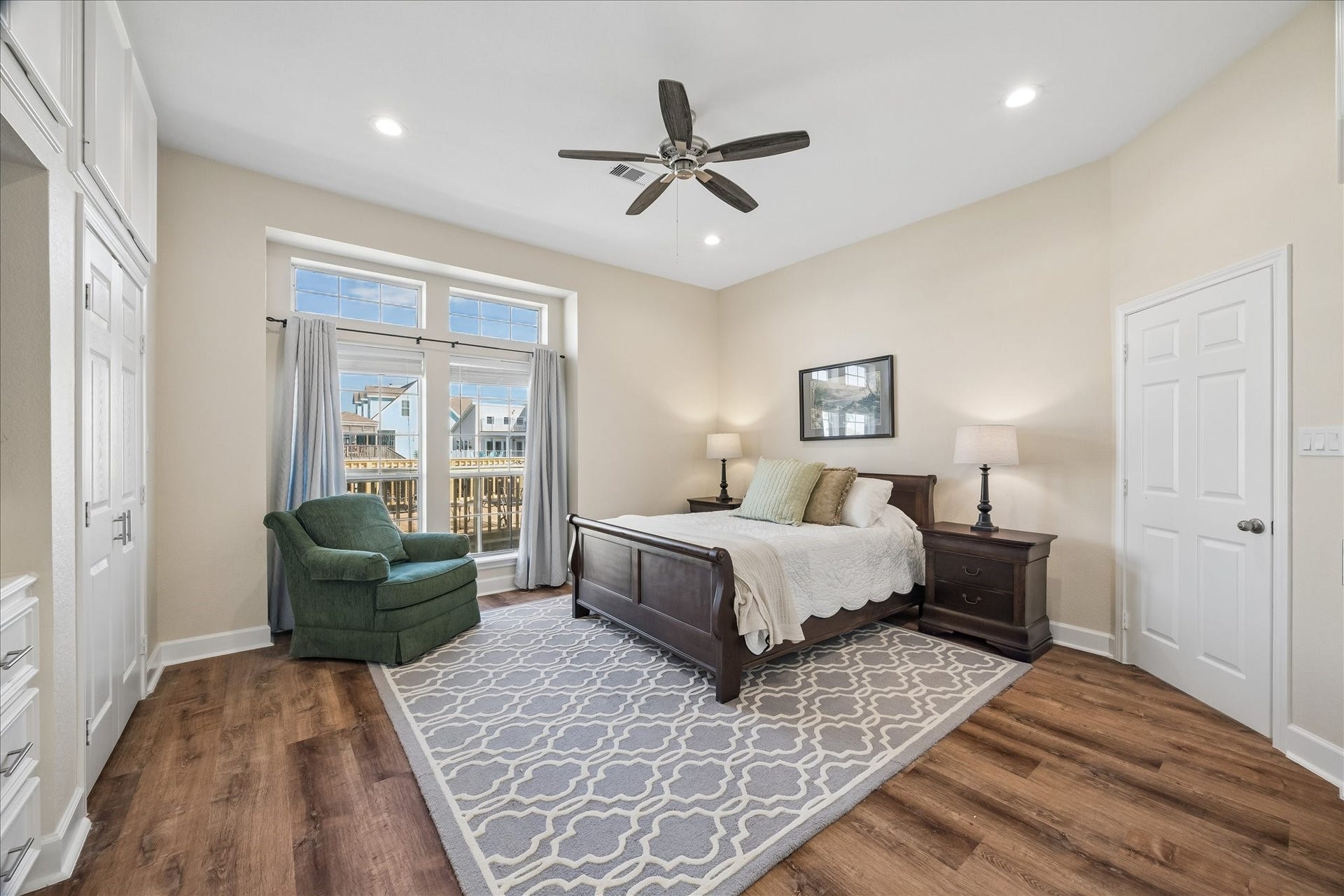 Bright and spacious bedroom with wood flooring, featuring a large bed, cozy green armchair, and elegant decor. Natural light floods in through the ample windows, complemented by a ceiling fan and recessed lighting.