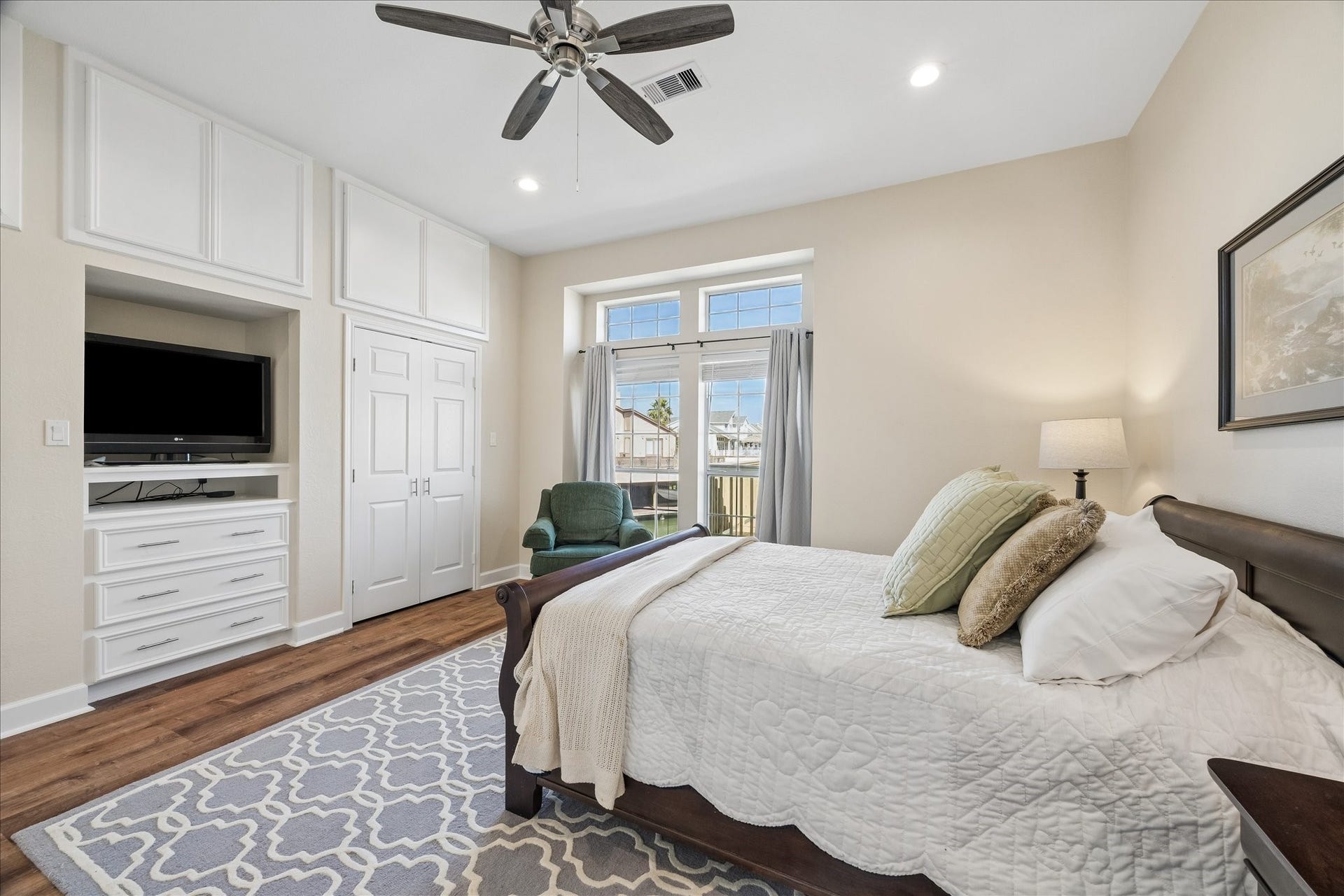 This cozy bedroom features a large window for natural light, a ceiling fan, built-in storage, a TV, and hardwood floors. It includes a comfortable bed with stylish bedding and a tasteful area rug, creating a welcoming atmosphere.