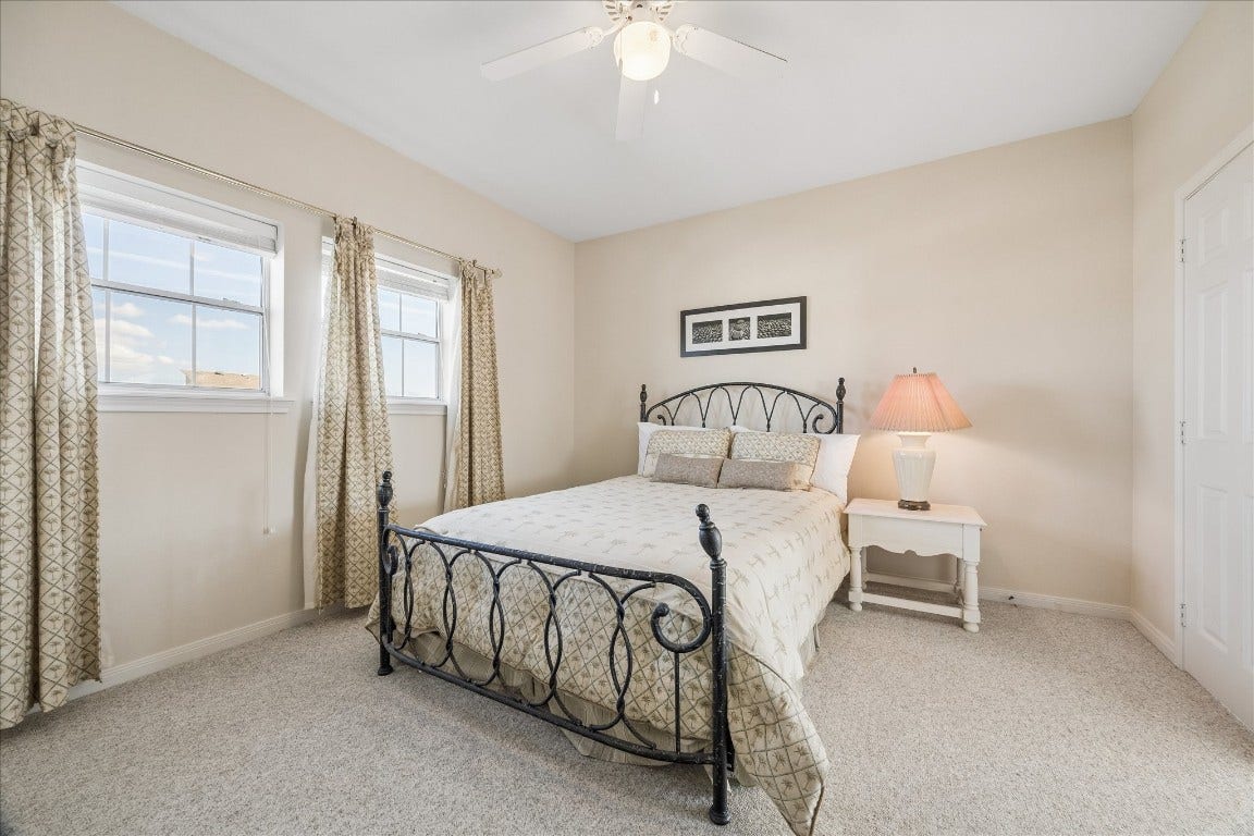 This cozy bedroom features a wrought iron bed, neutral walls, and carpeted flooring. Two windows with patterned curtains provide natural light, and a ceiling fan adds comfort. A bedside table with a lamp completes the inviting space.
