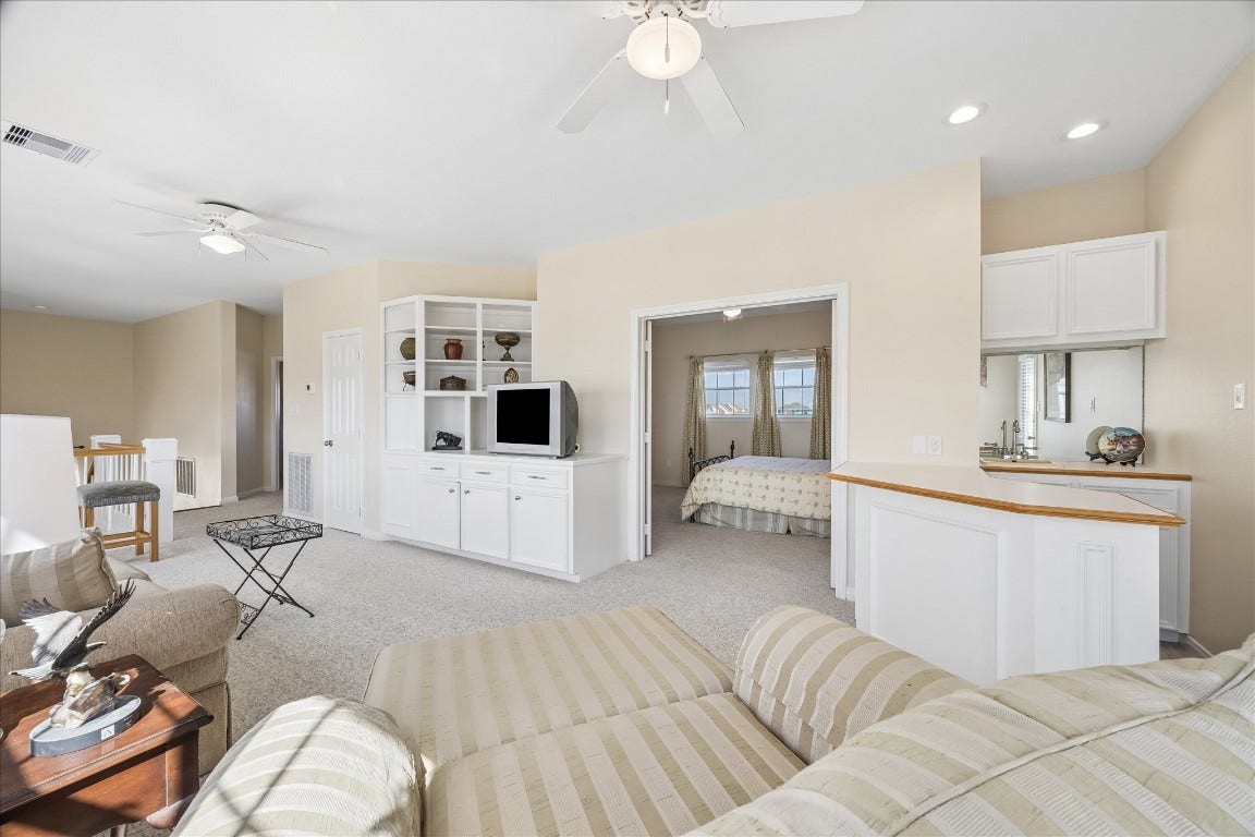This bright and spacious room features a cozy sitting area with a built-in entertainment center, open shelving, and a wet bar. A doorway leads to a bedroom with large windows, enhancing the natural light. Neutral tones and ceiling fans create a welcoming atmosphere.