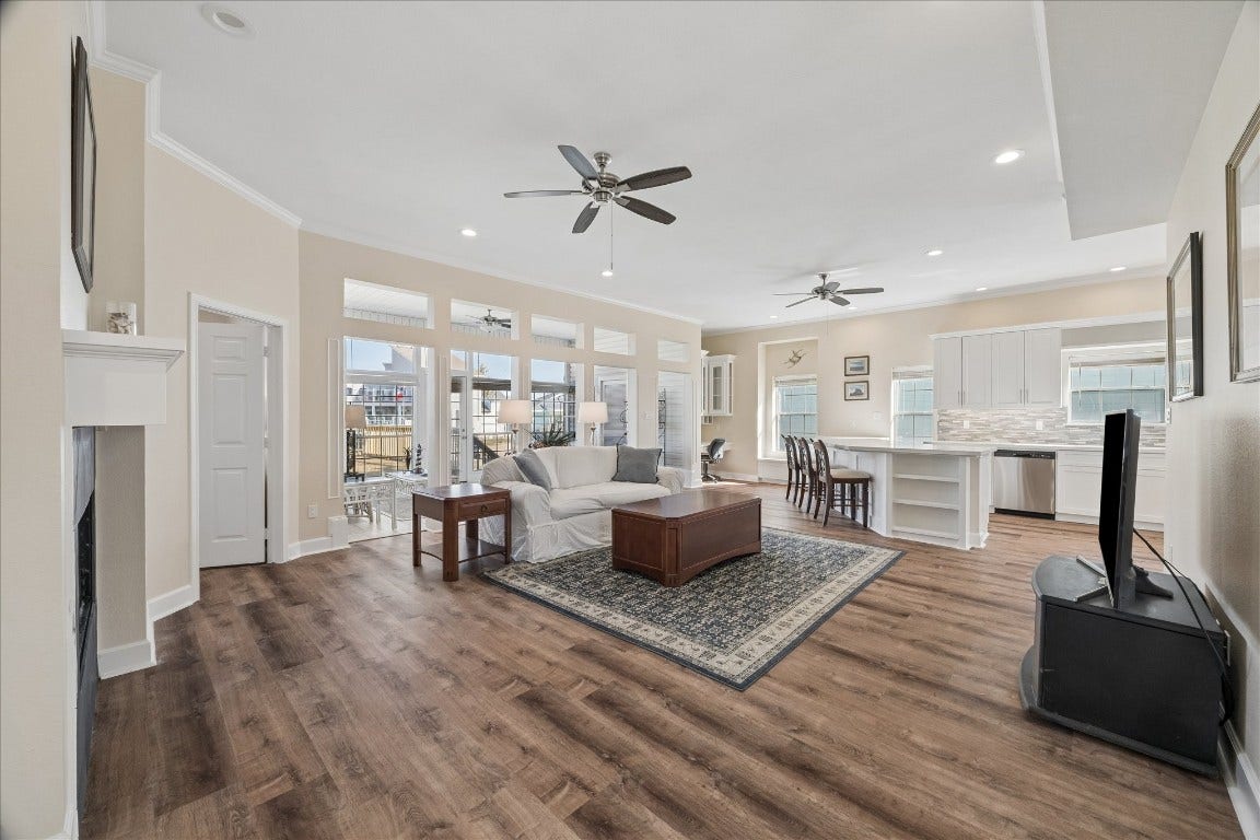 This spacious open-concept living area features elegant wood flooring and abundant natural light. It includes a cozy seating area with a fireplace, a modern kitchen with white cabinetry and a large island, and a dining space, perfect for entertaining. Large windows and ceiling fans enhance the bright and airy atmosphere.