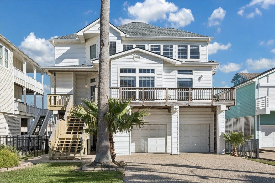 This is a charming, multi-story coastal home with a white exterior, featuring expansive windows and a large front deck. It has a double garage and is elevated with a stairway entrance, complemented by palm trees and a well-maintained front yard.