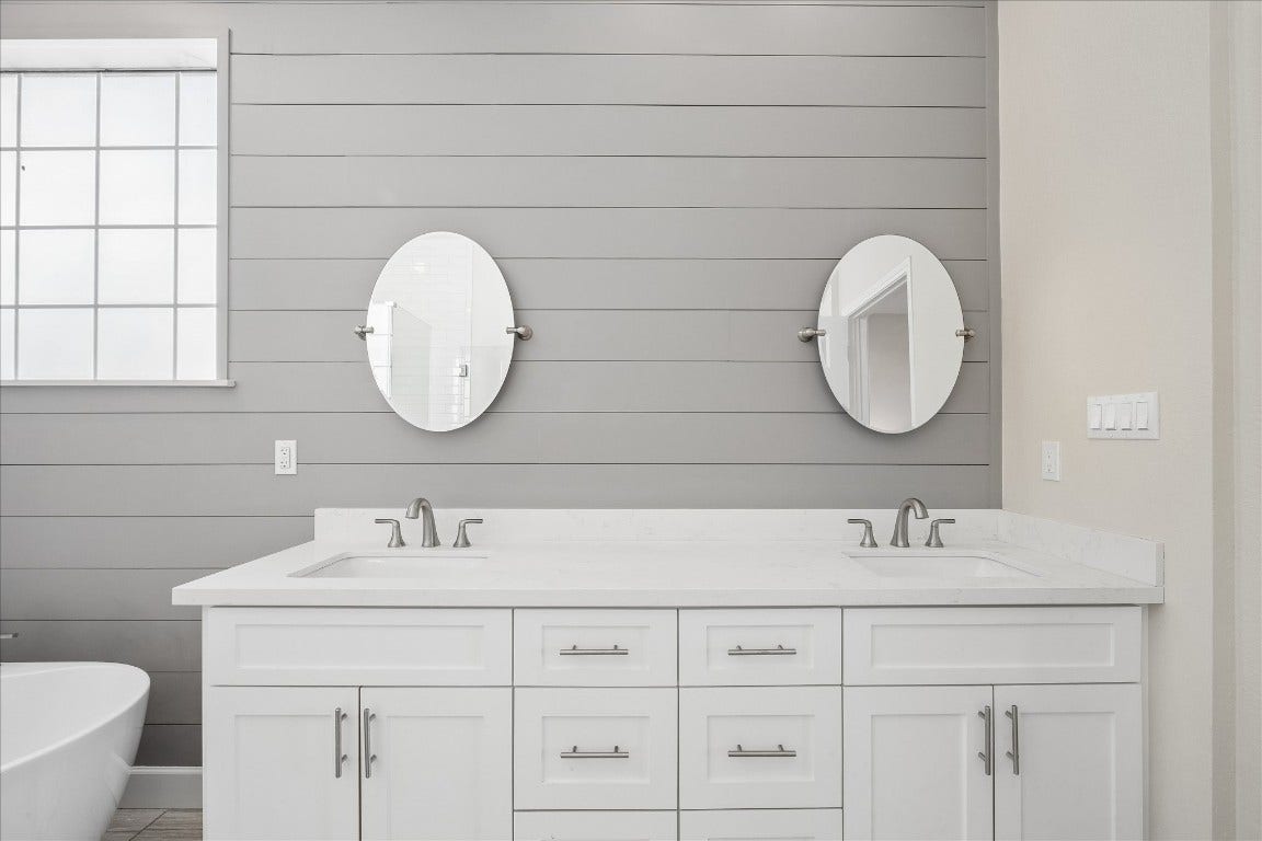 Modern bathroom features a double vanity with white cabinetry and sleek faucets, set against a gray shiplap wall. Two round mirrors and a window provide ample light, creating a bright, inviting space.