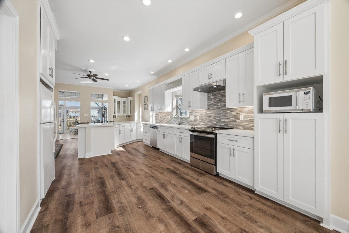 This spacious kitchen features modern white cabinetry, stainless steel appliances, and a stylish tile backsplash. It has a light, open layout with wood-style flooring, ample counter space, and abundant natural light.