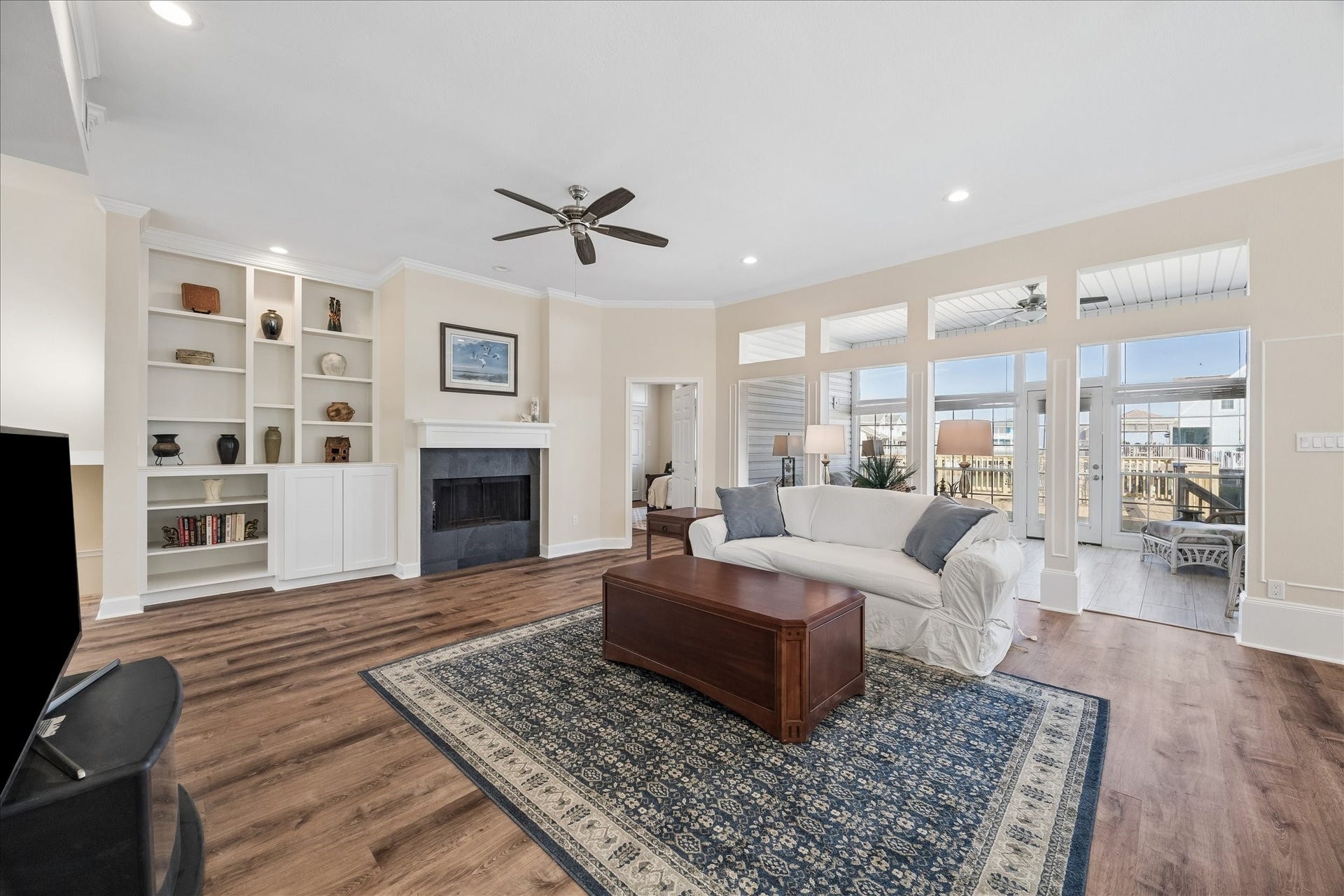 This spacious living room features a cozy fireplace with built-in shelving, large windows allowing plenty of natural light, and stylish wood flooring. A comfortable seating area and neutral tones create an inviting atmosphere, perfect for relaxation and entertaining.
