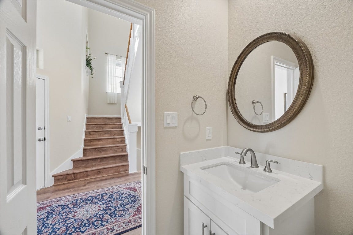 This photo features a bright, welcoming entryway with a staircase leading up, accented by a decorative rug. Adjacent is a small bathroom with a sleek vanity and an elegant round mirror. The space is well-lit and offers a modern, inviting feel.