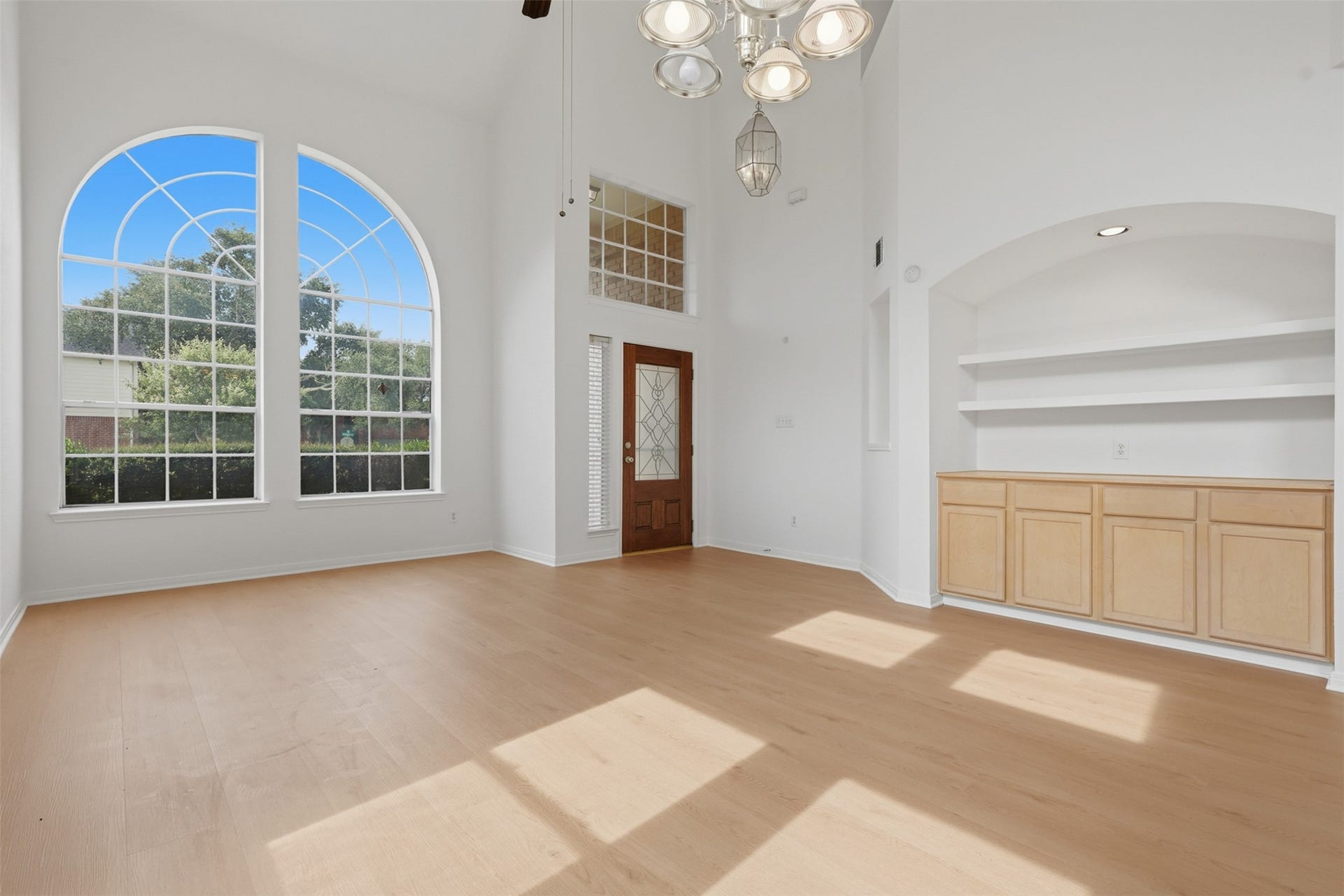 16622 Stoneside Drive, Houston, TX 77095 Double-height arched windows fill this spacious room with natural light, showcasing a recessed alcove with built-in cabinetry and floating display shelves above smooth plank flooring.