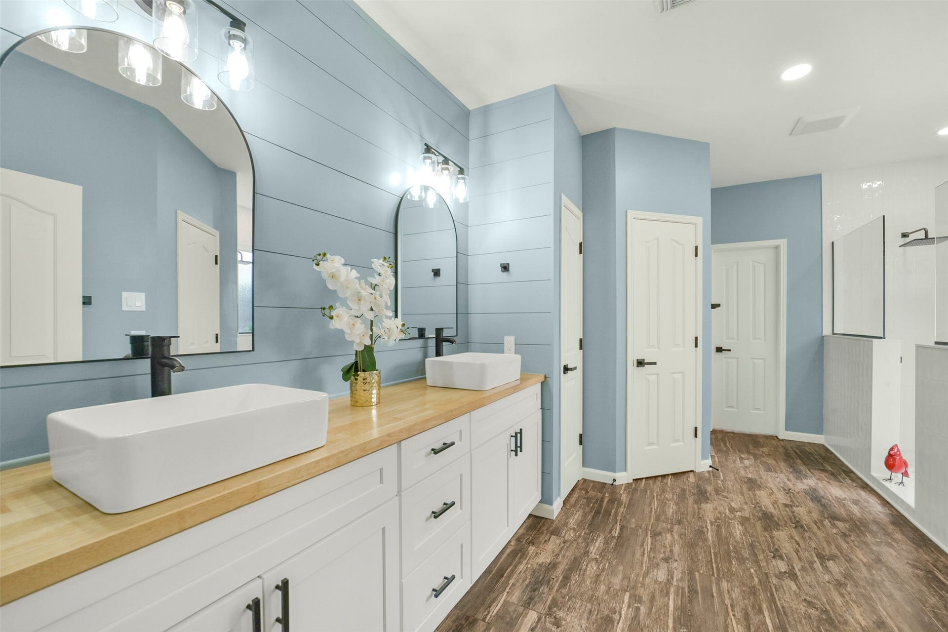 15730 Lake Loop Drive, Cypress, TX 77433 Elegant bathroom featuring dual vessel sinks on a sleek wooden countertop, complemented by modern black fixtures and stylish lighting. The blue shiplap walls and wood-look flooring create a serene, spa-like ambiance.