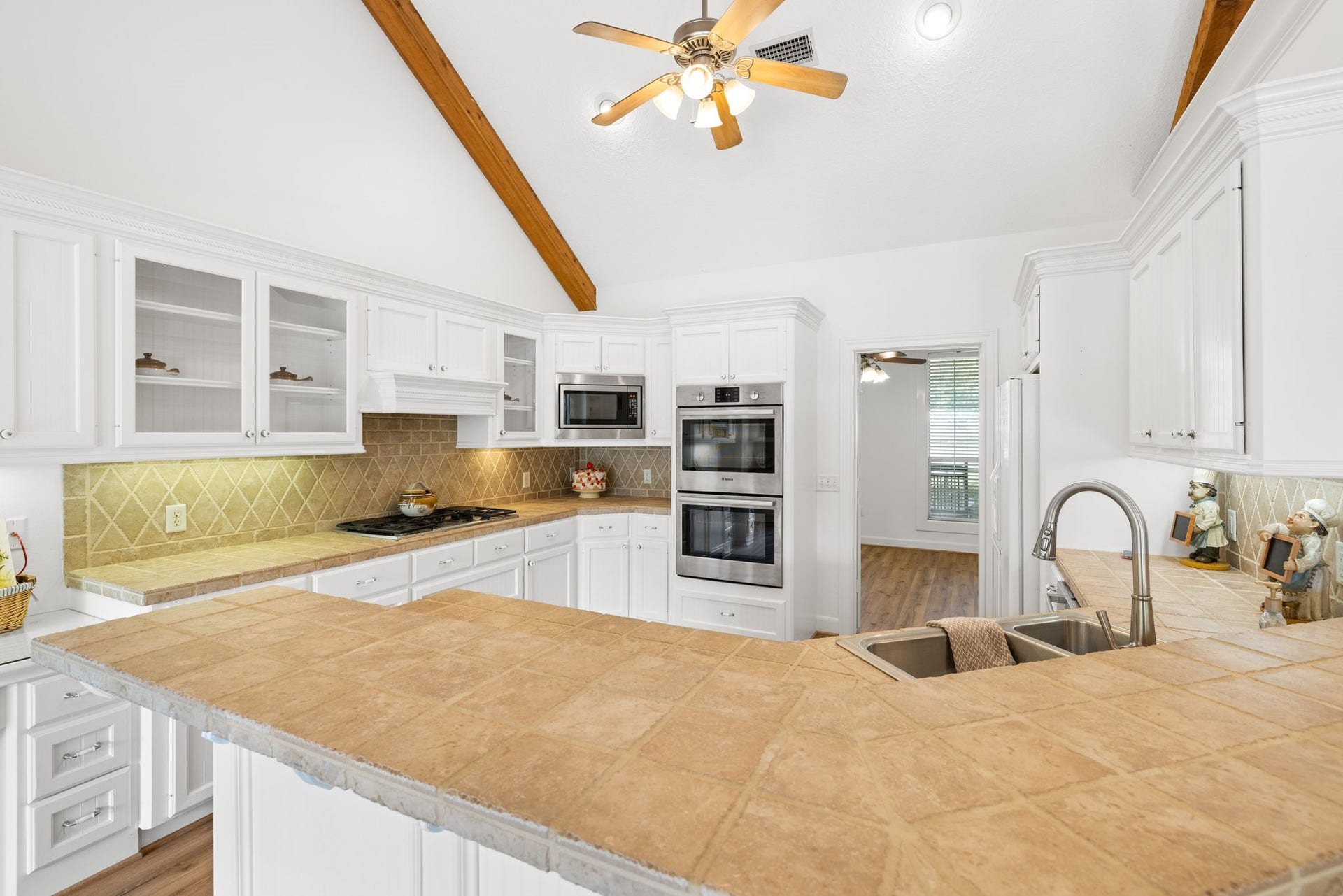 34310 High Point Drive, Magnolia, TX 77355 This kitchen features high vaulted ceilings with exposed wooden beams, white cabinetry, and a central island with a breakfast bar. It includes stainless steel appliances, a double oven, and a modern sink, all complemented by warm tile countertops and backsplash.