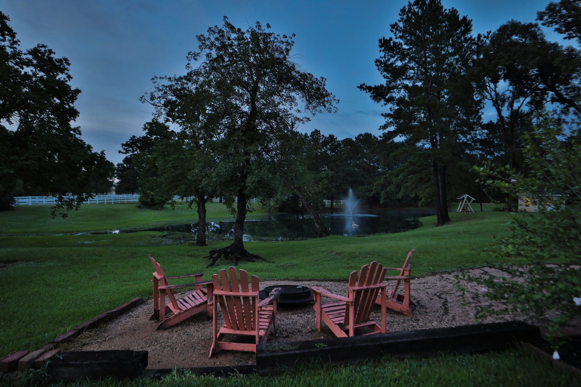 34310 High Point Drive, Magnolia, TX 77355 This photo showcases a serene property at dusk, featuring a spacious yard with mature trees, a tranquil pond with a fountain, and room for your cozy seating around a fire pit. Perfect place to unwind with your guests!