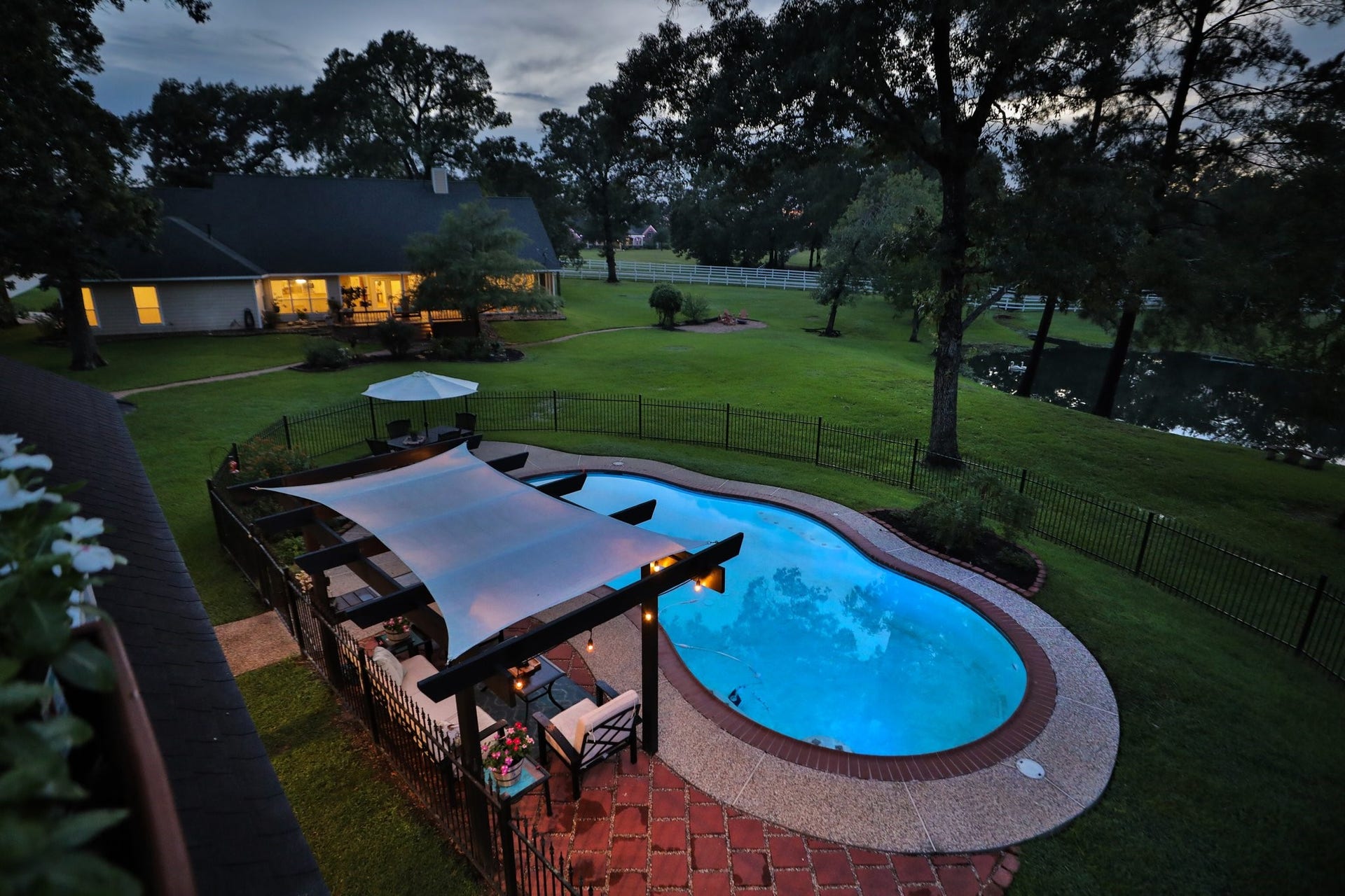 34310 High Point Drive, Magnolia, TX 77355 This aerial view shows a spacious backyard with a serene pool, shaded pergola area, and lush greenery, ideal for relaxation and entertaining. The house is warmly lit and offers a picturesque view of the pond and surrounding trees, creating a tranquil and inviting atmosphere.