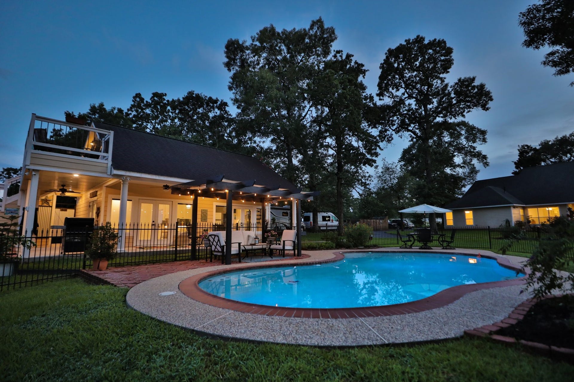 34310 High Point Drive, Magnolia, TX 77355 This photo showcases a charming backyard featuring a well-lit pool with surrounding patio space and guest house. The adjacent deck and seating area under a pergola offer a perfect spot for relaxation or entertaining. Lush greenery and mature trees provide a peaceful, private atmosphere, complemented by the warm glow from the home's exterior lighting.