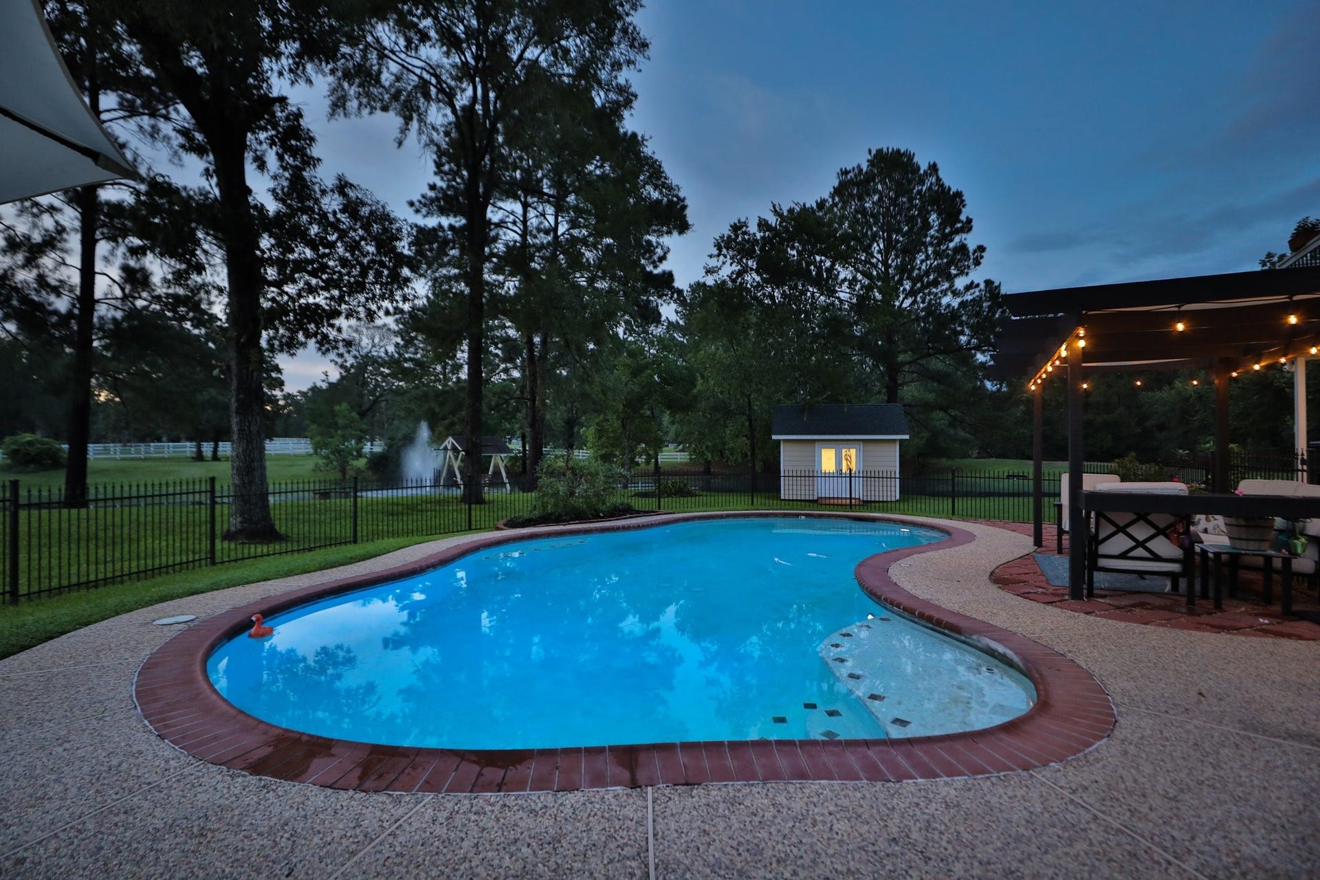 34310 High Point Drive, Magnolia, TX 77355 Sunset view of this inviting backyard features a well-lit swimming pool surrounded by lush greenery and a fenced-in area for privacy. A cozy pergola with seating area offers a perfect spot for relaxing or entertaining.