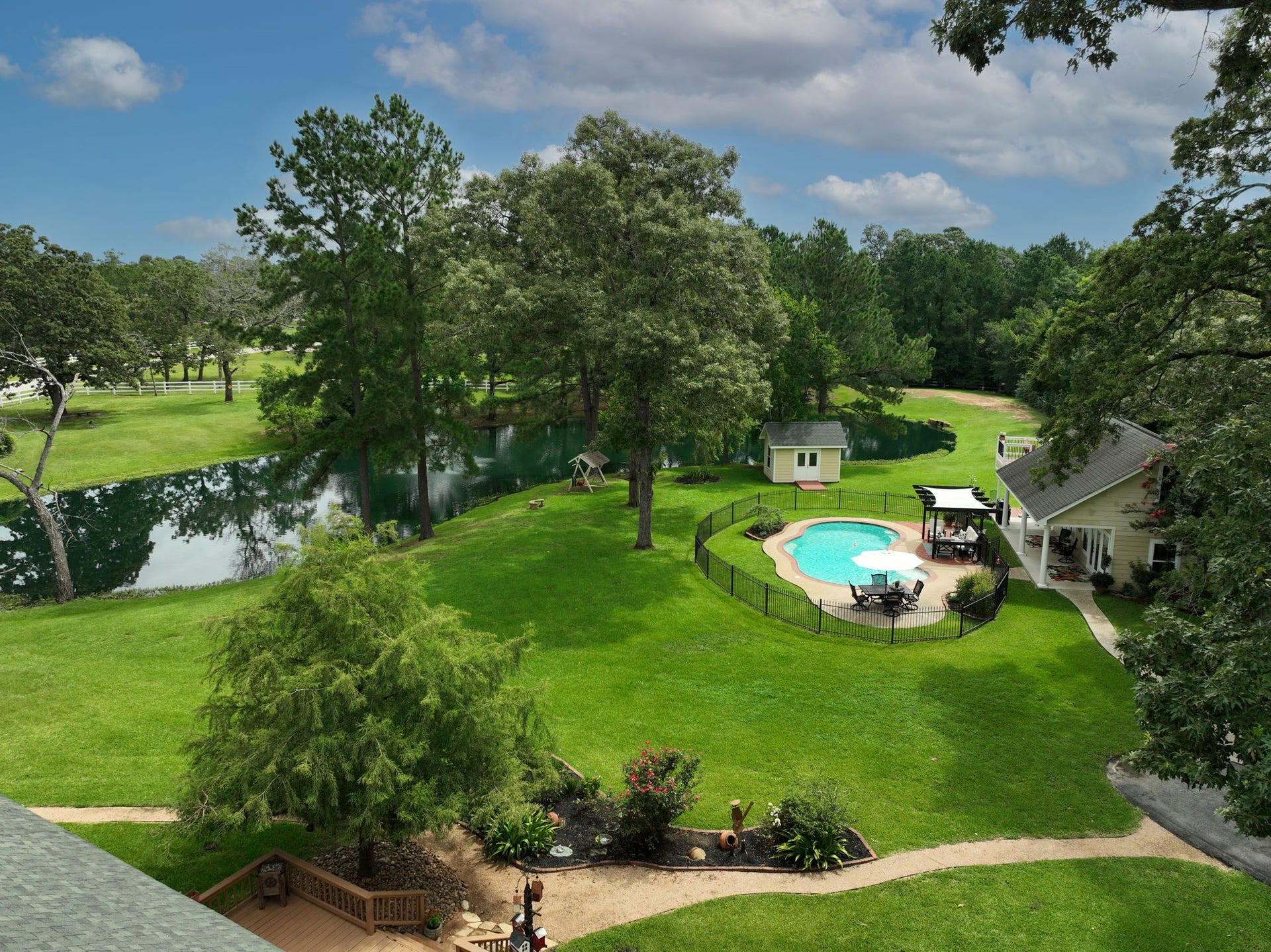 34310 High Point Drive, Magnolia, TX 77355 This aerial view shows lush greenery, a serene pond, and a fenced-in pool area. The charming guest house with a pergola offers a perfect spot for relaxation, surrounded by mature trees and manicured landscaping. Ideal for those seeking a peaceful retreat.
