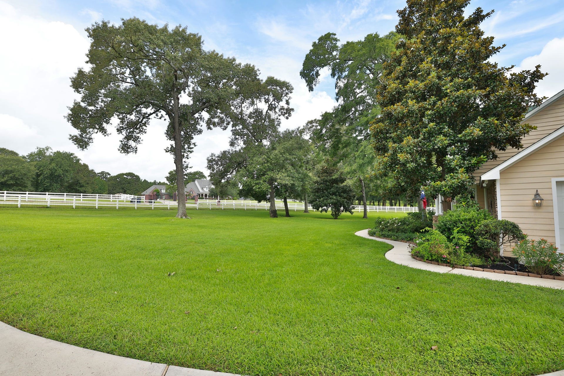 34310 High Point Drive, Magnolia, TX 77355 This photo showcases a spacious, well-maintained yard with lush green grass and mature trees, bordered by a white heavy gage dog/predator fence. The side of a house is visible, neatly landscaped and a curved walkway, suggesting a peaceful and inviting property.