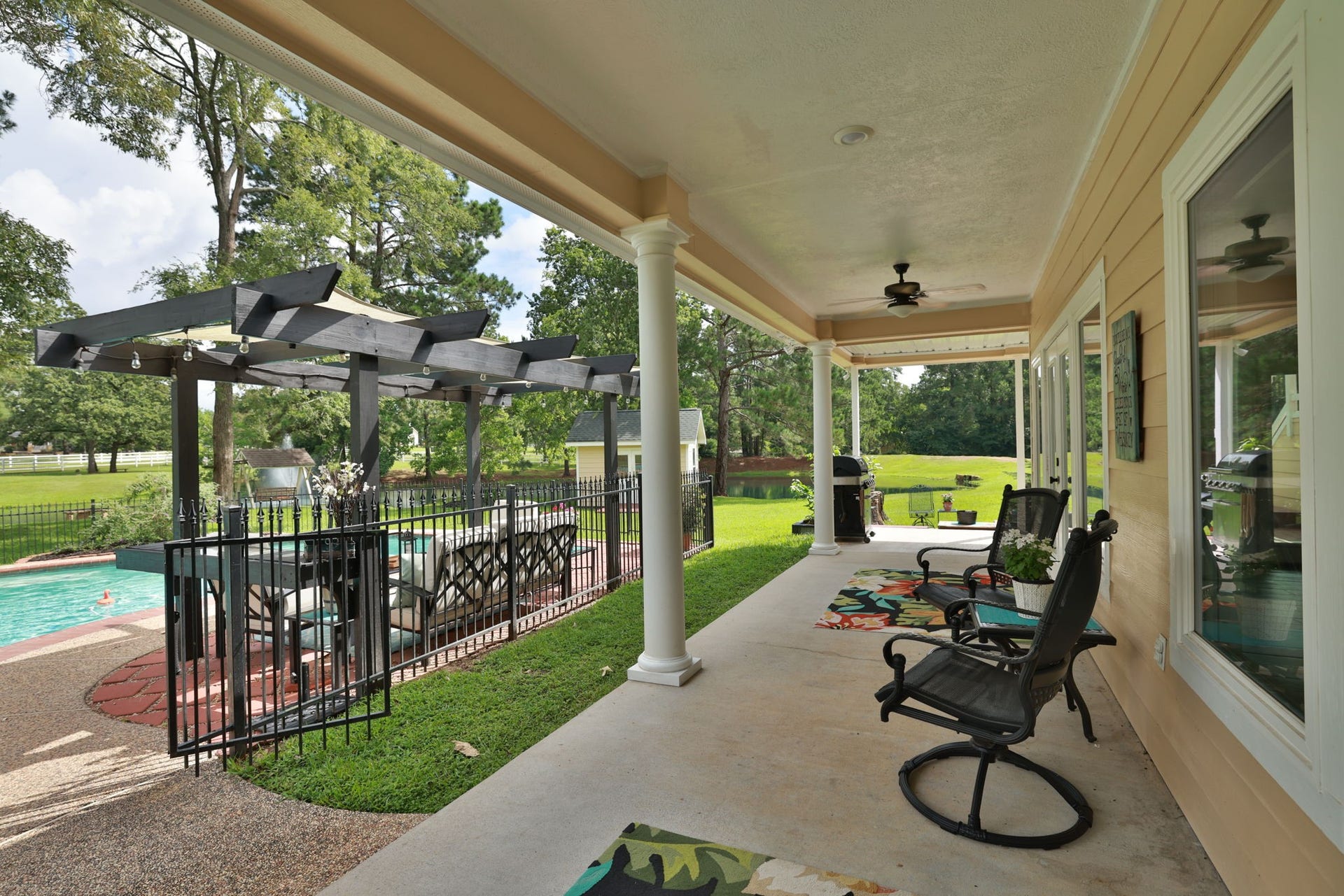 34310 High Point Drive, Magnolia, TX 77355 This photo showcases a spacious covered patio with ceiling fans, overlooking a fenced pool area with a pergola and seating. It offers a serene view of a lush green yard, perfect for outdoor relaxation and entertaining.