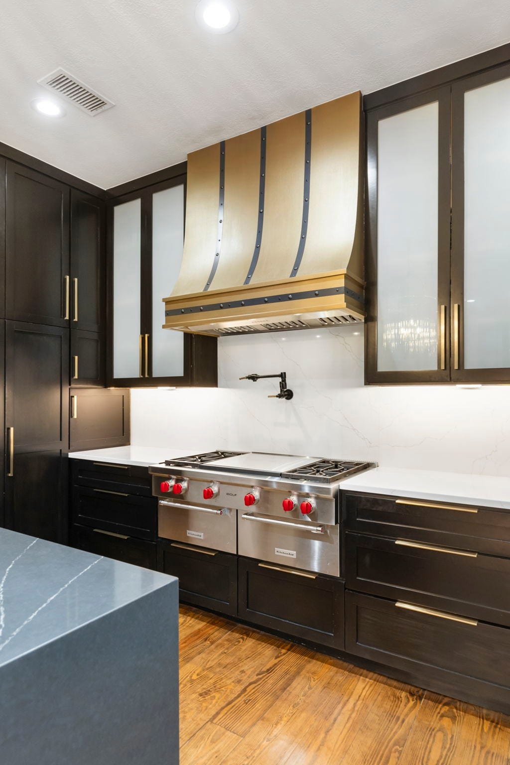 A statement brass & iron-strapped vent hood anchors the kitchen, set above a Wolf range with a pot filler.